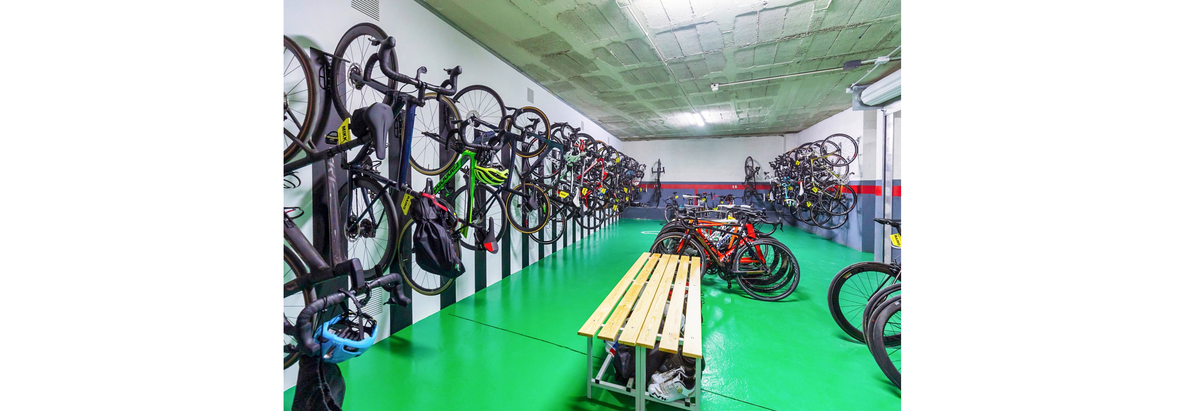 A brightly lit indoor room is filled with bicycles stored vertically on wall-mounted racks. Road and racing bikes line both sides of the space, with helmets and bags hanging from some frames. A long wooden bench stands in the center on a glossy green floor, providing a place to sit or prepare gear. The scene suggests an organized, secure bike parking area in an urban building or workplace.