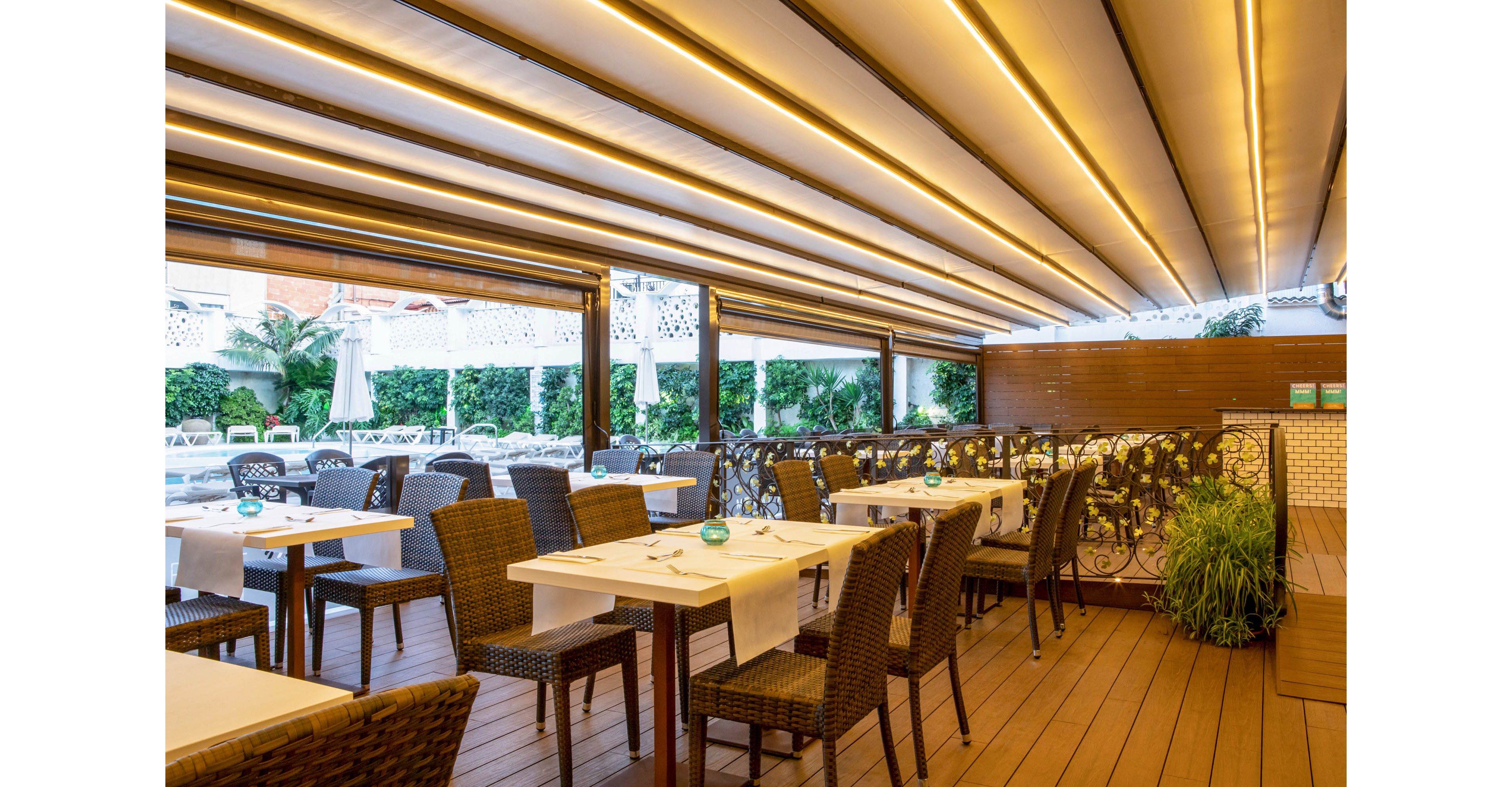 A stylish outdoor restaurant terrace features neatly arranged tables with white tablecloths and dark woven chairs. The space is covered by a modern retractable ceiling with warm linear lighting that creates a cozy ambiance. Green plants and hedges line the perimeter, adding a natural touch to the contemporary design. The setting appears calm and ready for guests, ideal for relaxed dining or evening gatherings.