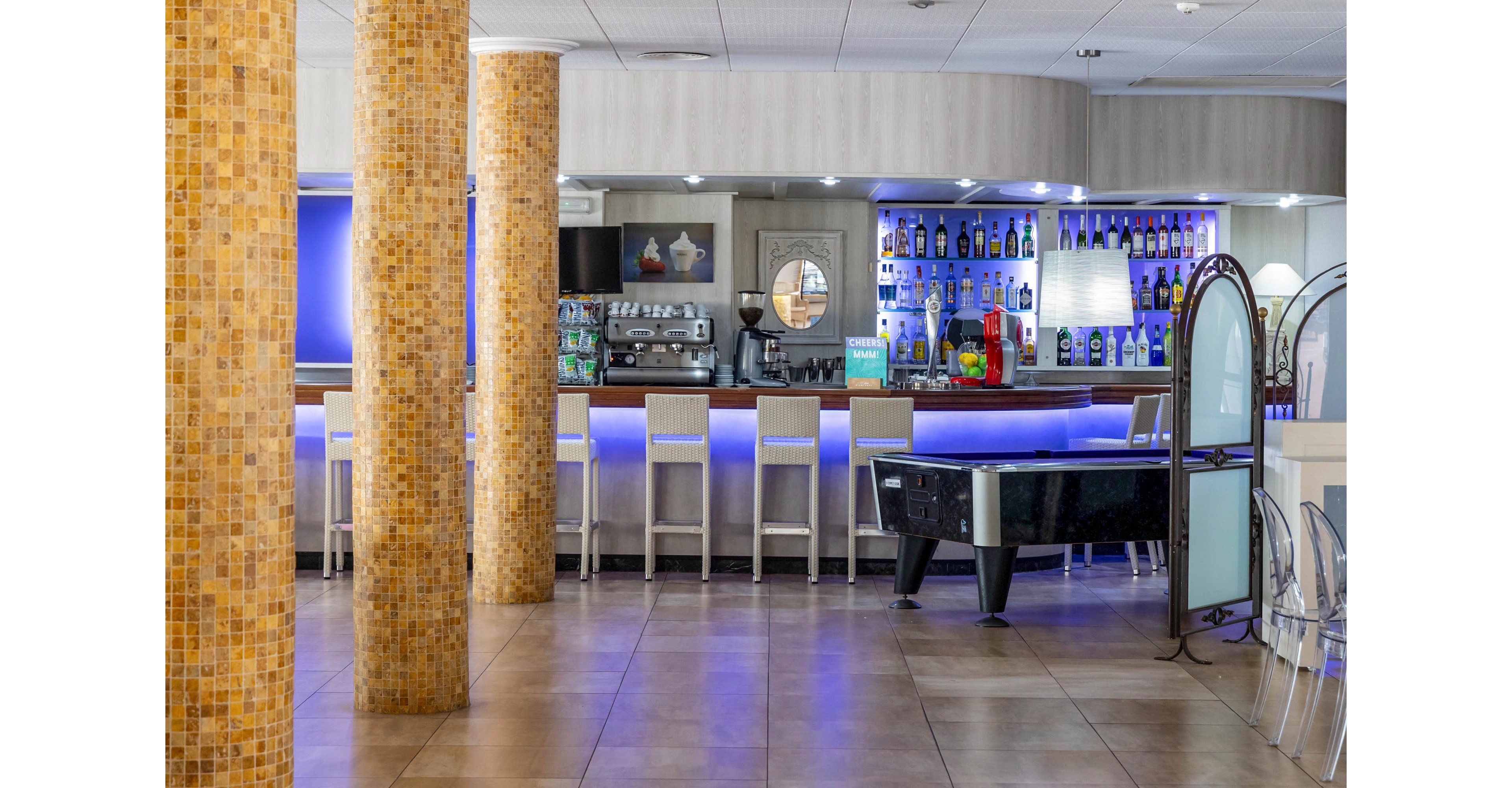 Contemporary bar interior featuring a long counter with illuminated blue backlighting and rows of bottles on display. High bar stools line the counter, while an air hockey or pool-style game table stands in the foreground. Mosaic-tiled columns and glossy floor tiles add a polished, modern look to the space. The setting appears quiet and empty, suggesting a relaxed lounge atmosphere.