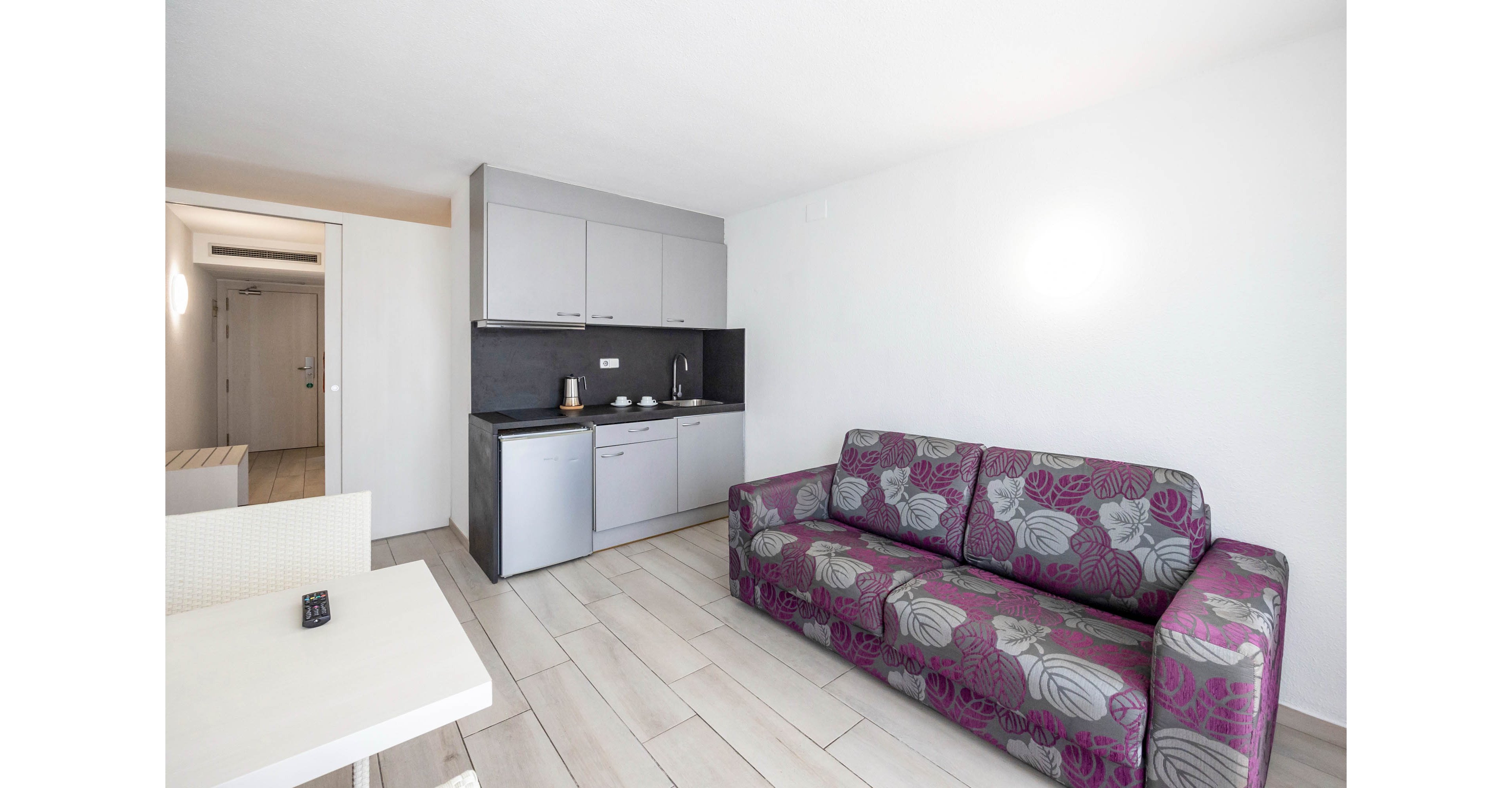 A bright studio apartment features a compact gray kitchenette with built-in cabinets and a small refrigerator. A purple patterned sofa sits against the wall, providing seating in the open-plan living area. Light wood-style flooring and white walls create a clean, minimalist look. A small dining table with a remote control on top is visible in the foreground, and a hallway leads to the entrance door in the background.