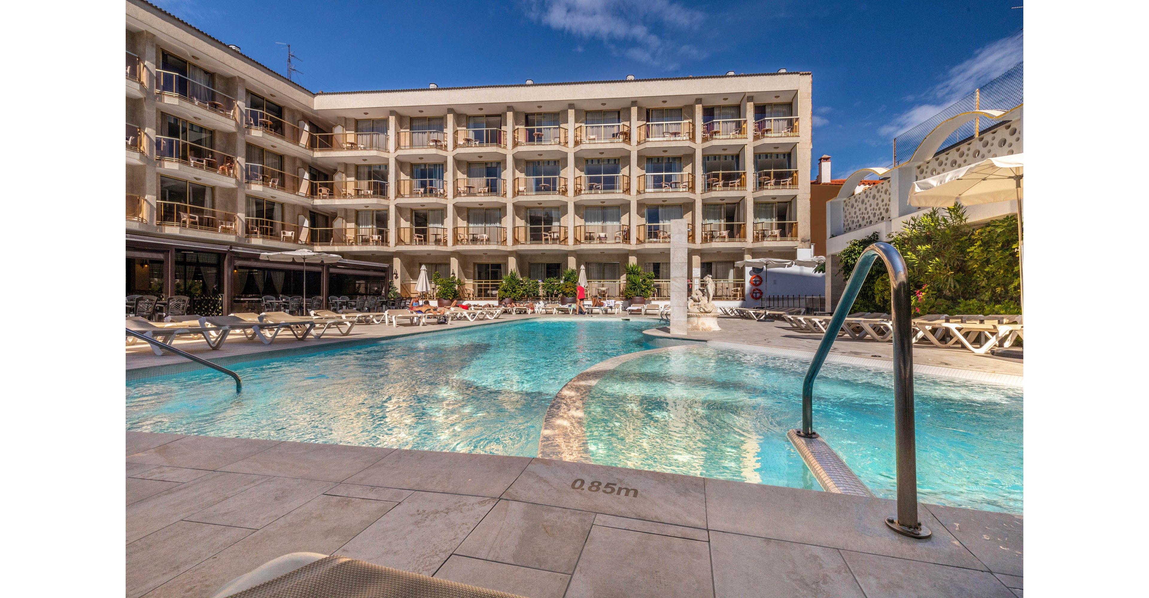 A large outdoor swimming pool sits in the courtyard of a multi-story hotel building. Sun loungers line the poolside terrace under a bright blue sky with scattered clouds. Stainless steel handrails lead into the clear water, and the pool depth marking of 0.85m is visible on the edge. The scene suggests a relaxing vacation atmosphere at a resort or holiday accommodation.
