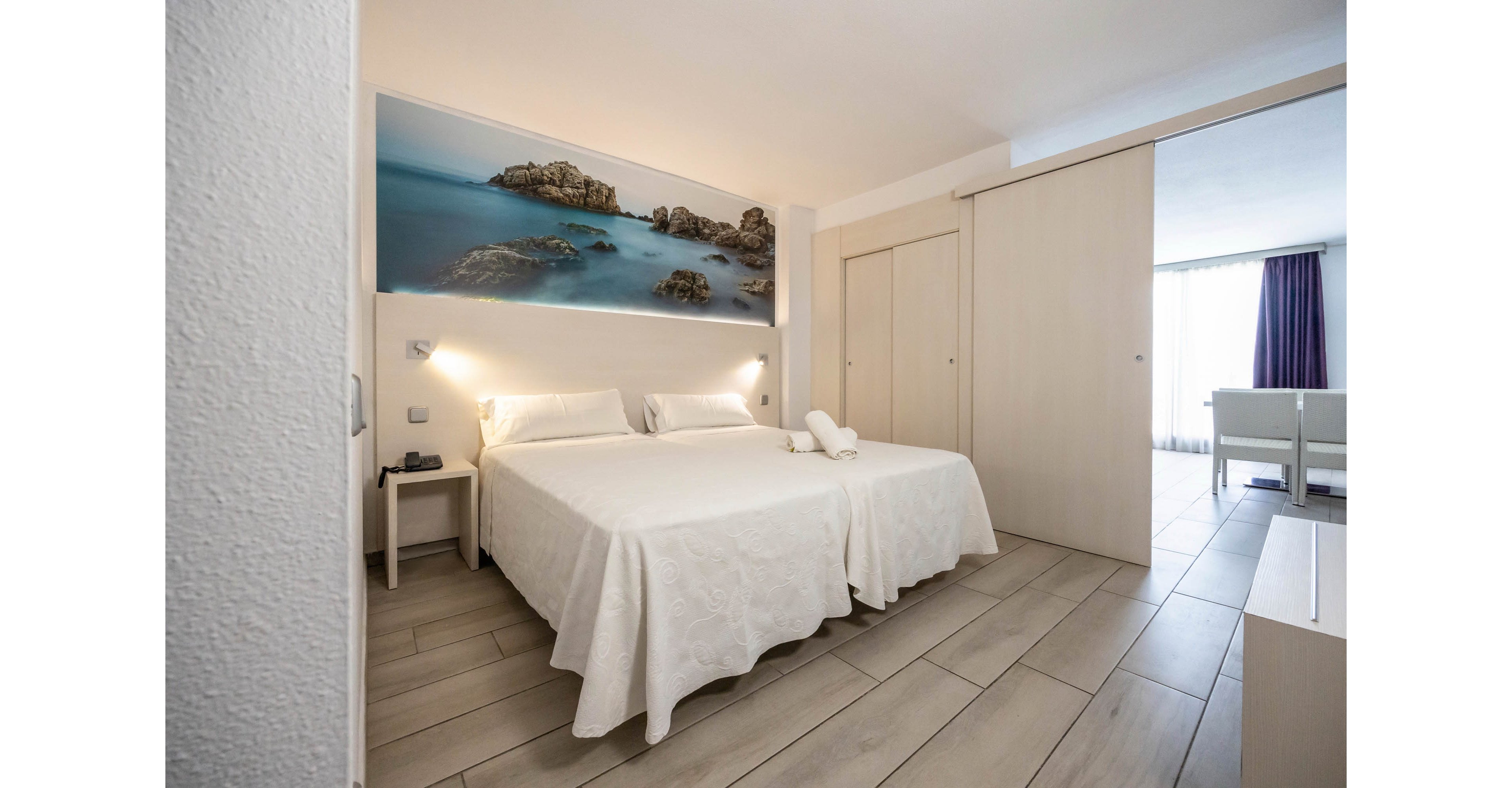 A bright, modern hotel room features two neatly made twin beds with white linens and folded towels placed on top. The space is decorated in neutral tones with light wood furniture and tiled flooring. A large wall art photograph of a rocky seaside landscape hangs above the beds, adding a coastal touch. Sliding doors open to a sitting area with a sofa and natural light streaming in through a window with curtains.