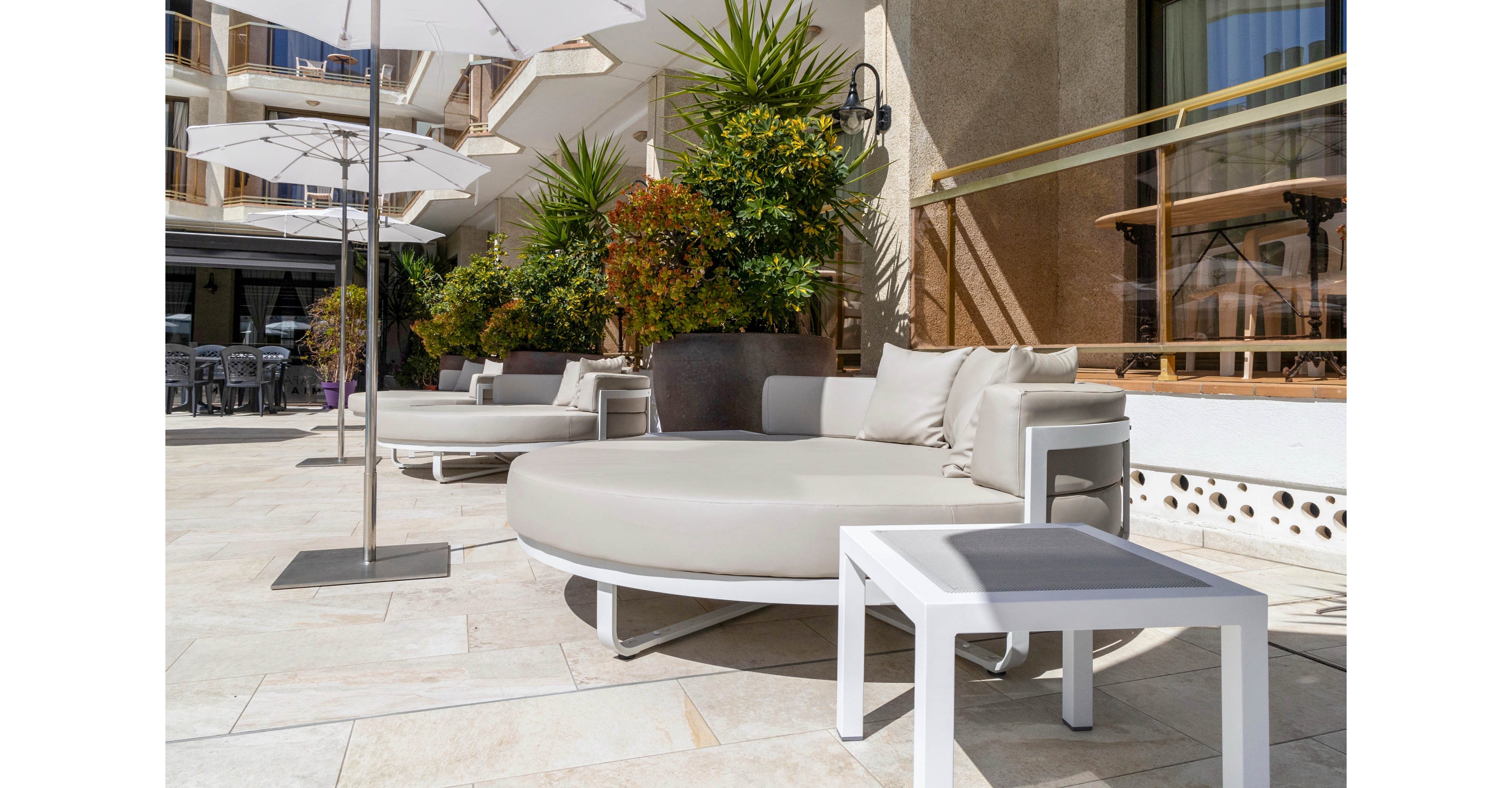 A contemporary outdoor lounge area features large round cushioned daybeds and a small white side table on a tiled terrace. White umbrellas provide shade while potted plants and greenery add a touch of nature along the building facade. The scene is brightly lit by sunlight, highlighting the neutral tones of the furniture and flooring. The setting suggests a relaxing hotel or resort patio designed for comfort and leisure.