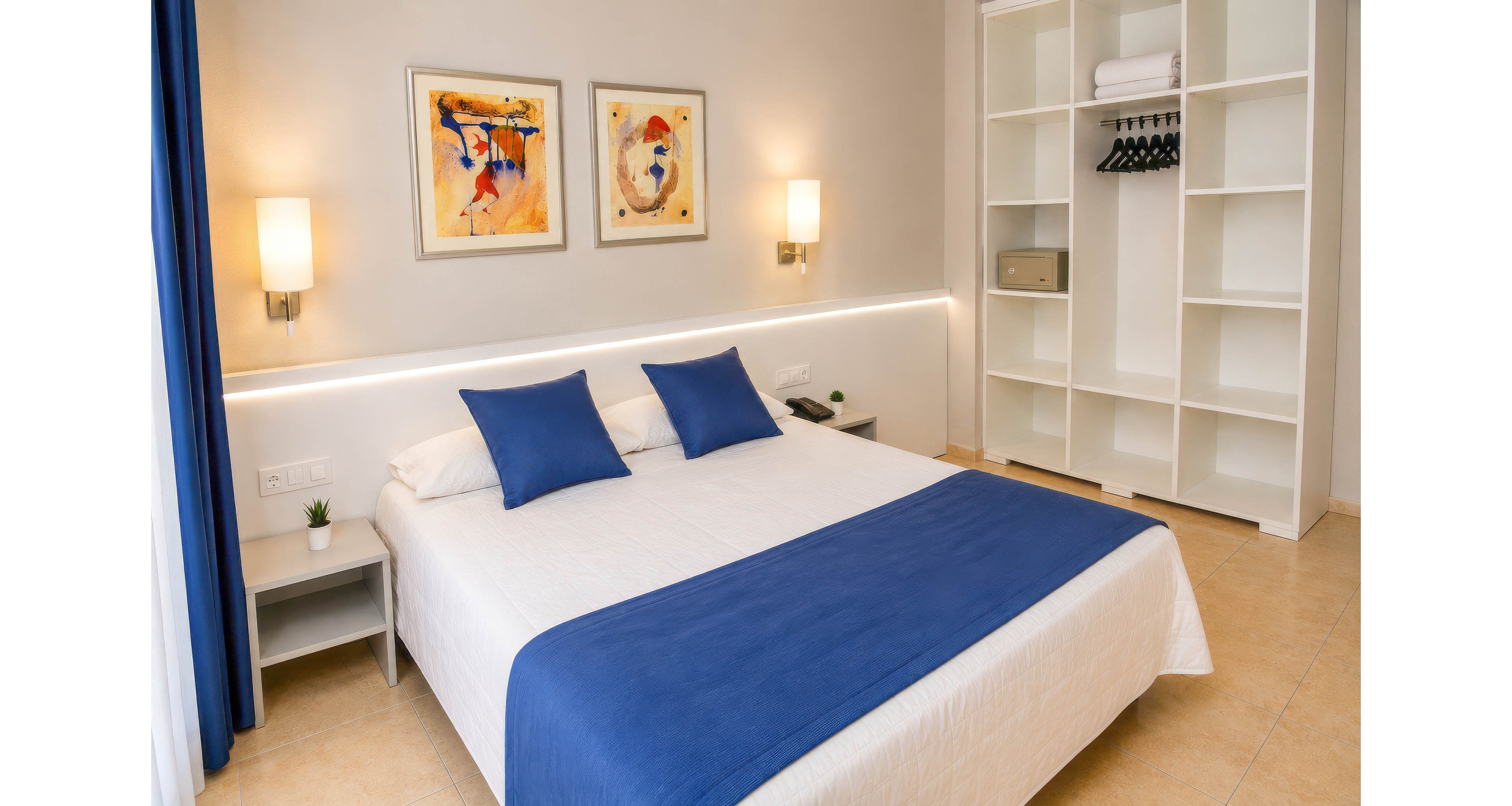 Bright, contemporary bedroom featuring a double bed with white linens and blue pillows and runner. The room includes minimalist bedside tables, wall-mounted lamps, and abstract artwork above the bed. An open wardrobe with hangers and shelves is visible, adding to the clean, organized look. Neutral walls and flooring contrast with the bold blue textiles and curtain, creating a fresh, inviting atmosphere.