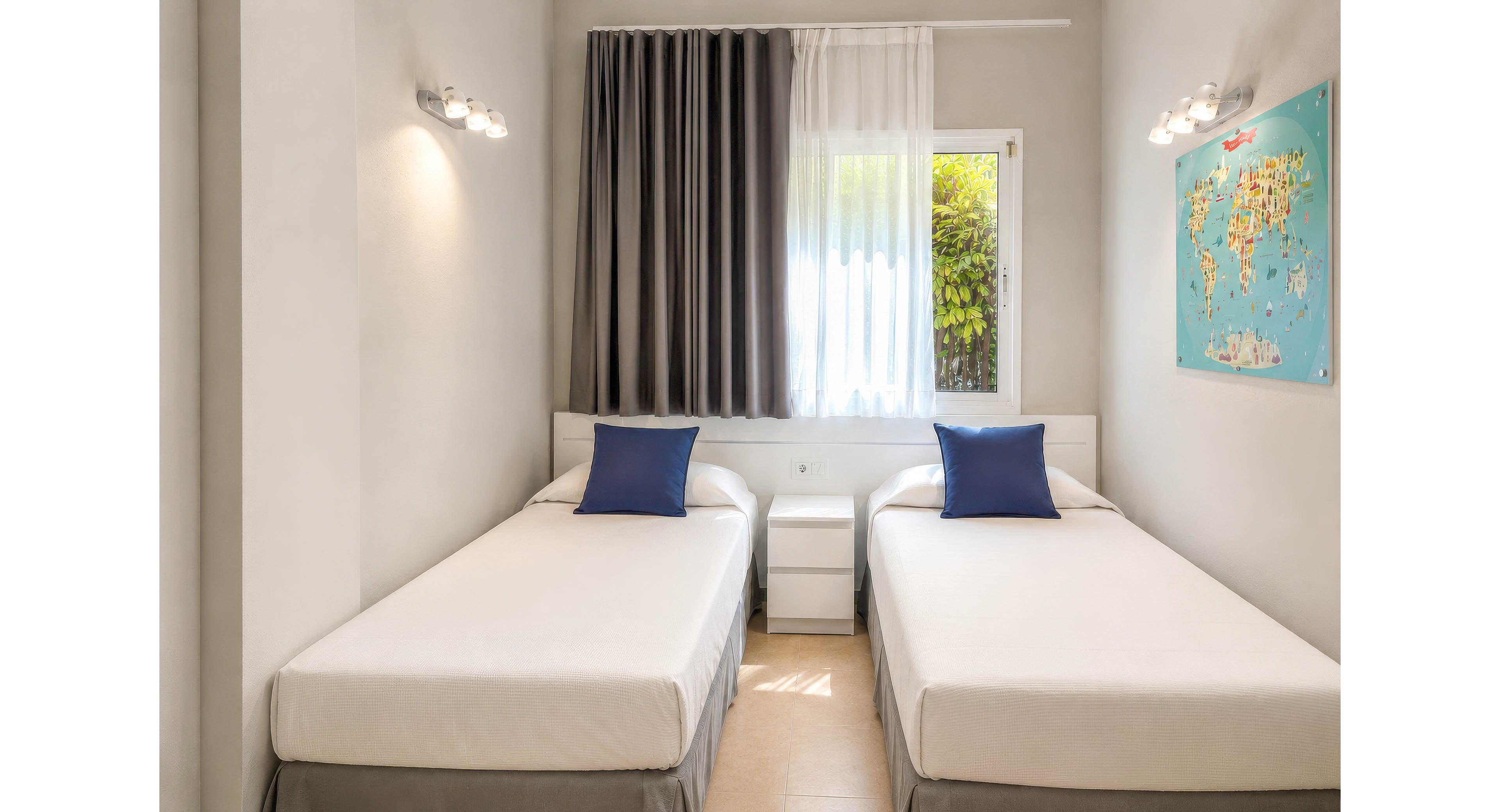 A clean, modern bedroom features two single beds separated by a small white nightstand. Each bed has a white duvet and a blue accent pillow, creating a simple, fresh look. Natural light enters through a window with sheer white curtains and dark gray drapes, revealing greenery outside. A colorful world map artwork hangs on the wall, adding a decorative touch to the neutral space.