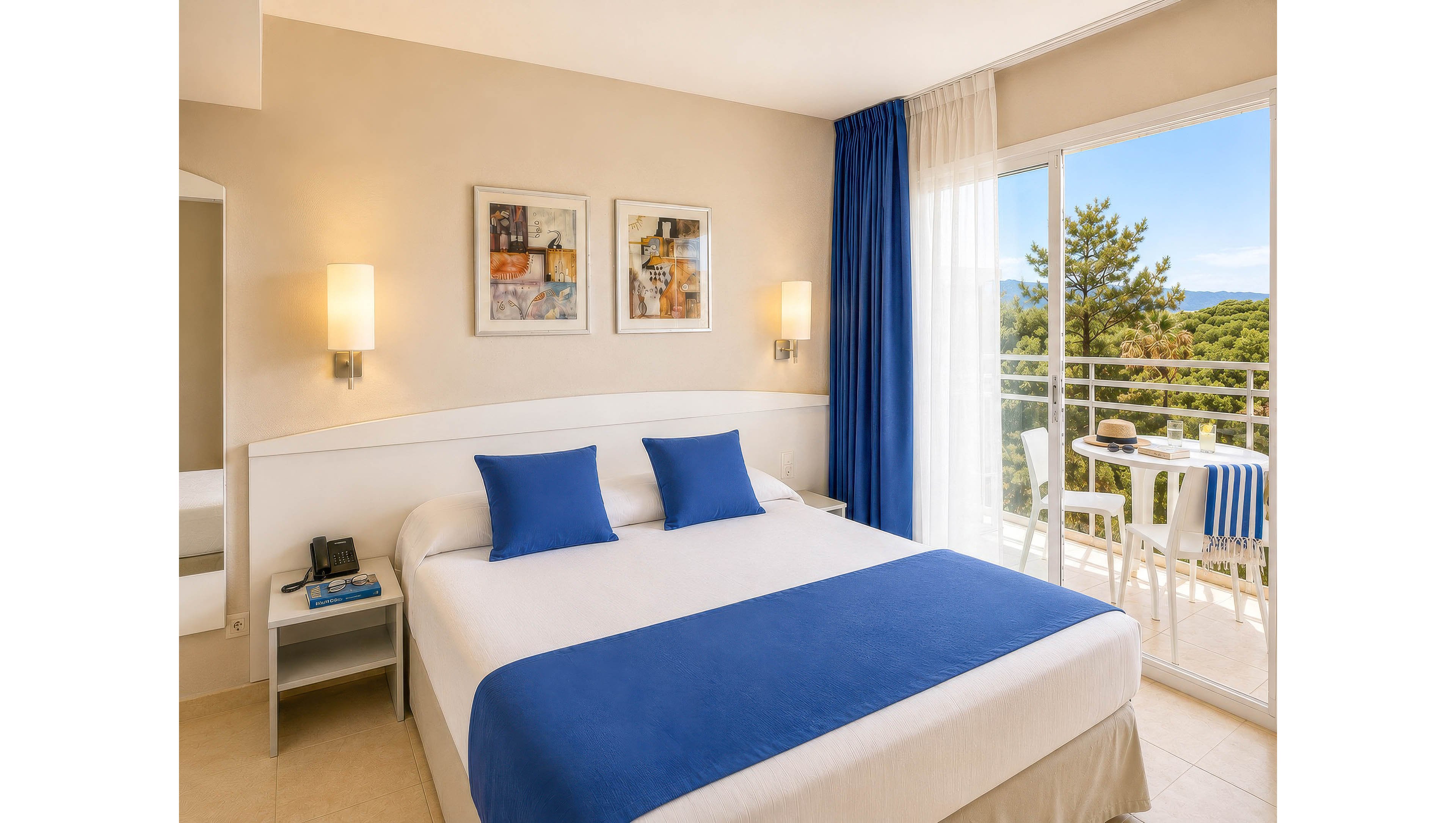 Modern hotel room featuring a large double bed with blue pillows and a matching blue bed runner. The space is decorated in neutral tones with two wall-mounted lamps and abstract artwork above the headboard. A glass door opens to a balcony with white furniture and a striped blue and white tablecloth, overlooking greenery and distant hills. Natural daylight fills the room, creating a clean and inviting atmosphere.