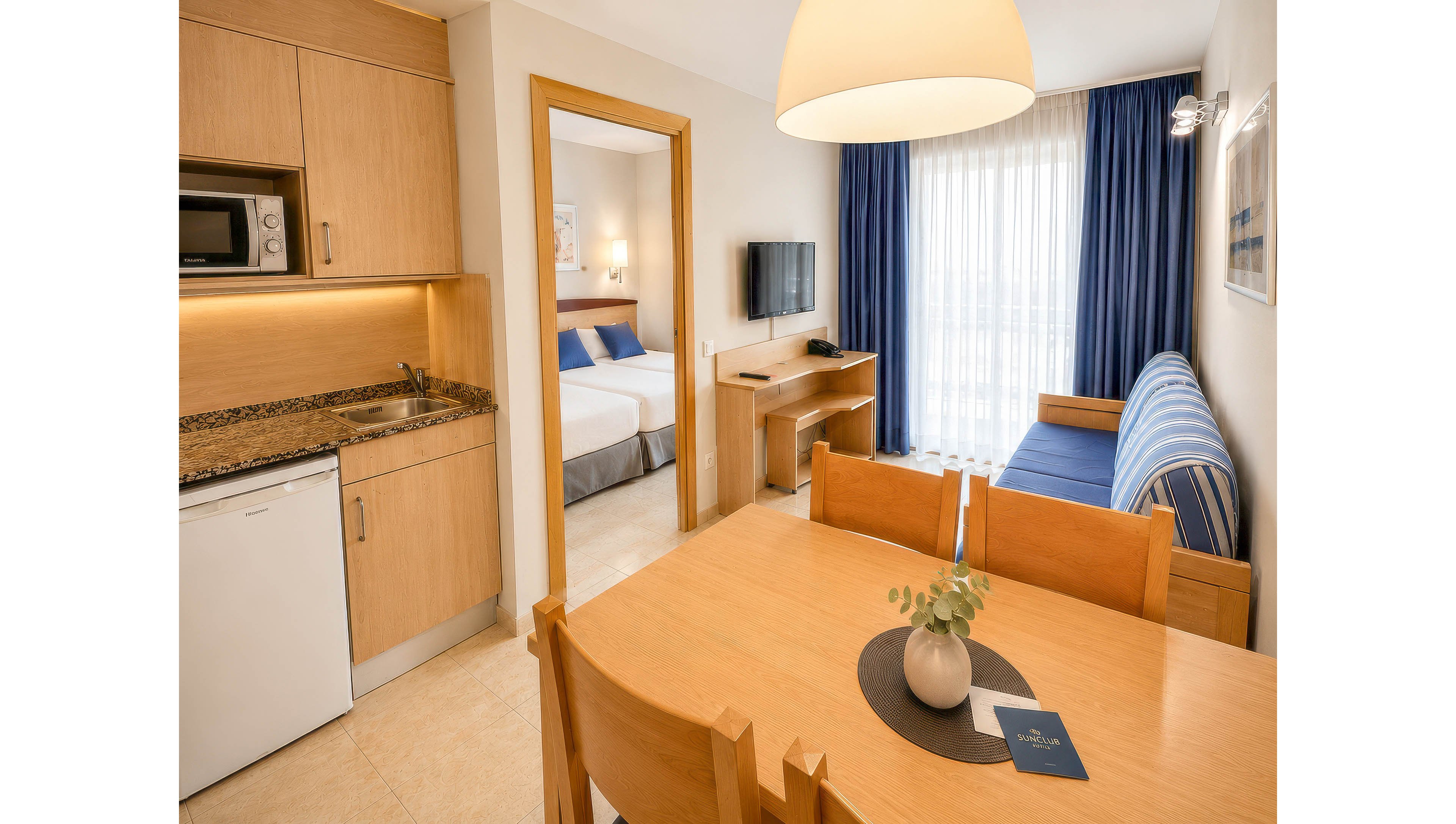 A modern hotel suite features a small kitchenette with wooden cabinets, a microwave, and a compact refrigerator. In the foreground, a wooden dining table with chairs holds a decorative vase and a booklet. The living area includes a striped blue sofa, a wall-mounted television, and a desk positioned near large windows with sheer curtains and blue drapes. Through an open doorway, a bedroom with a neatly made bed and blue accent pillows is visible, creating a clean and inviting atmosphere.