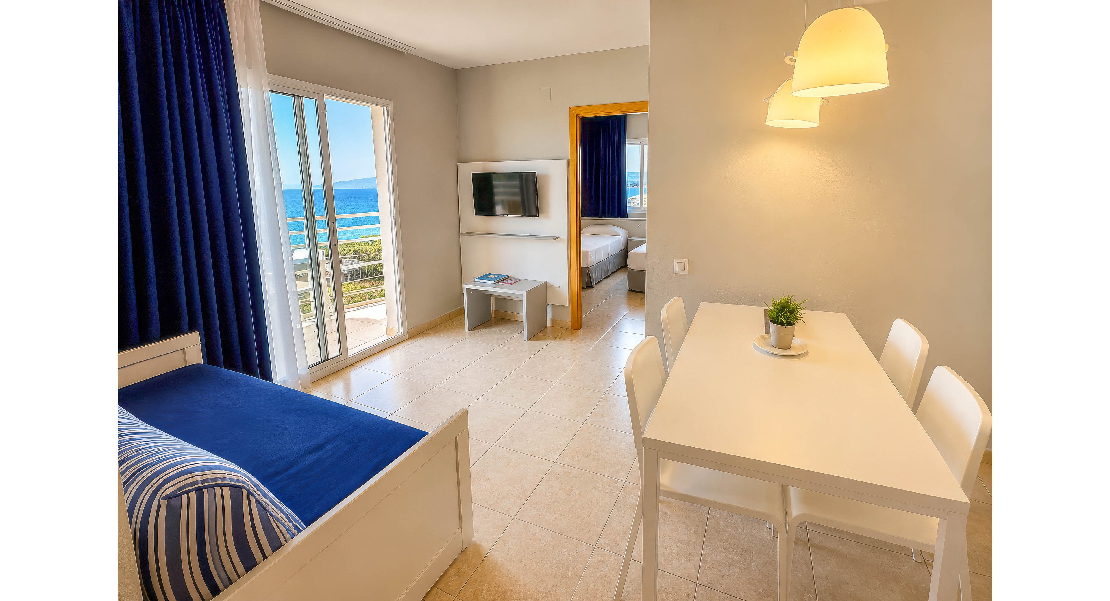 Modern hotel room interior featuring a white dining table and chairs beside a blue-upholstered daybed. Large glass doors open to a balcony with a clear view of the ocean and coastline. A wall-mounted television and small table sit near the doorway leading to a separate bedroom. Warm lighting and blue-and-white decor create a clean, inviting seaside atmosphere.