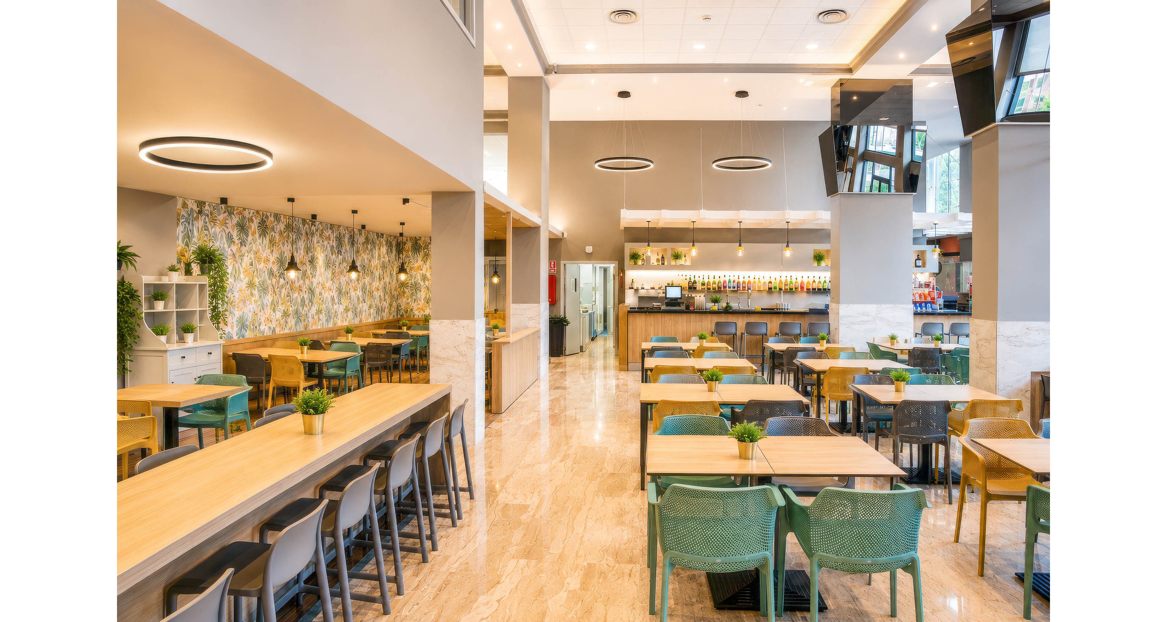 Bright, contemporary cafeteria interior featuring rows of wooden tables and mixed-color chairs. The space includes a long communal table, smaller square tables, and green chairs with potted plants as centerpieces. A patterned accent wall, ceiling ring lights, and marble-style flooring create a clean, modern atmosphere. The back counter area displays a variety of drinks and food items under warm lighting, with large windows bringing in natural light.
