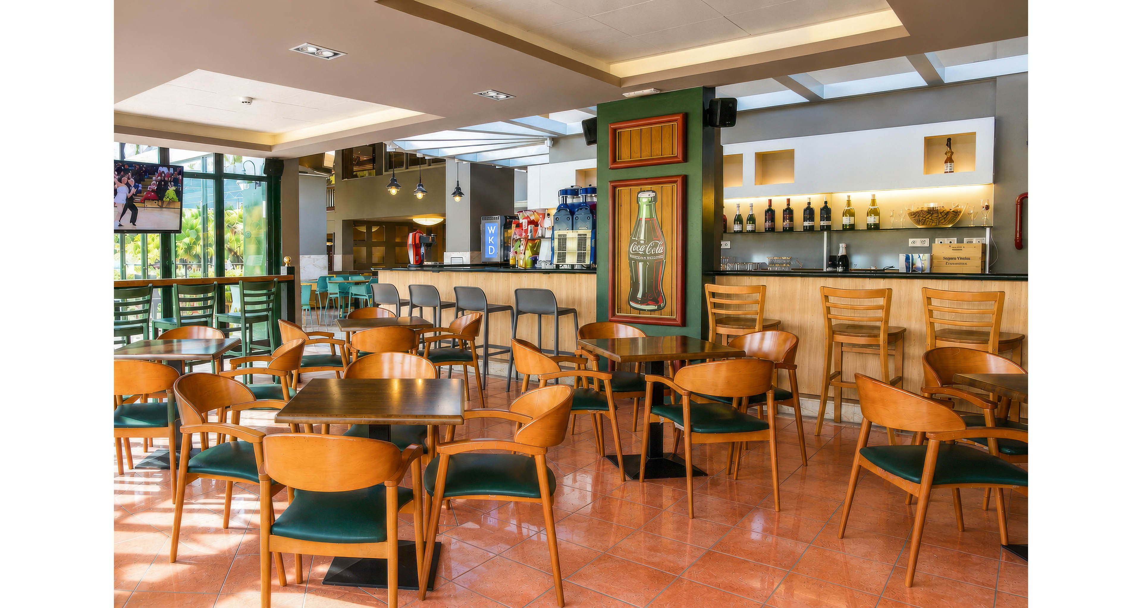A spacious café bar interior features numerous wooden tables and chairs neatly arranged on a tiled floor. The bar counter at the back displays bottles, glassware, and drink dispensers under warm lighting. Large windows on the side bring in natural daylight and offer views of greenery outside. Wall art and a television screen add visual interest to the relaxed, contemporary atmosphere.