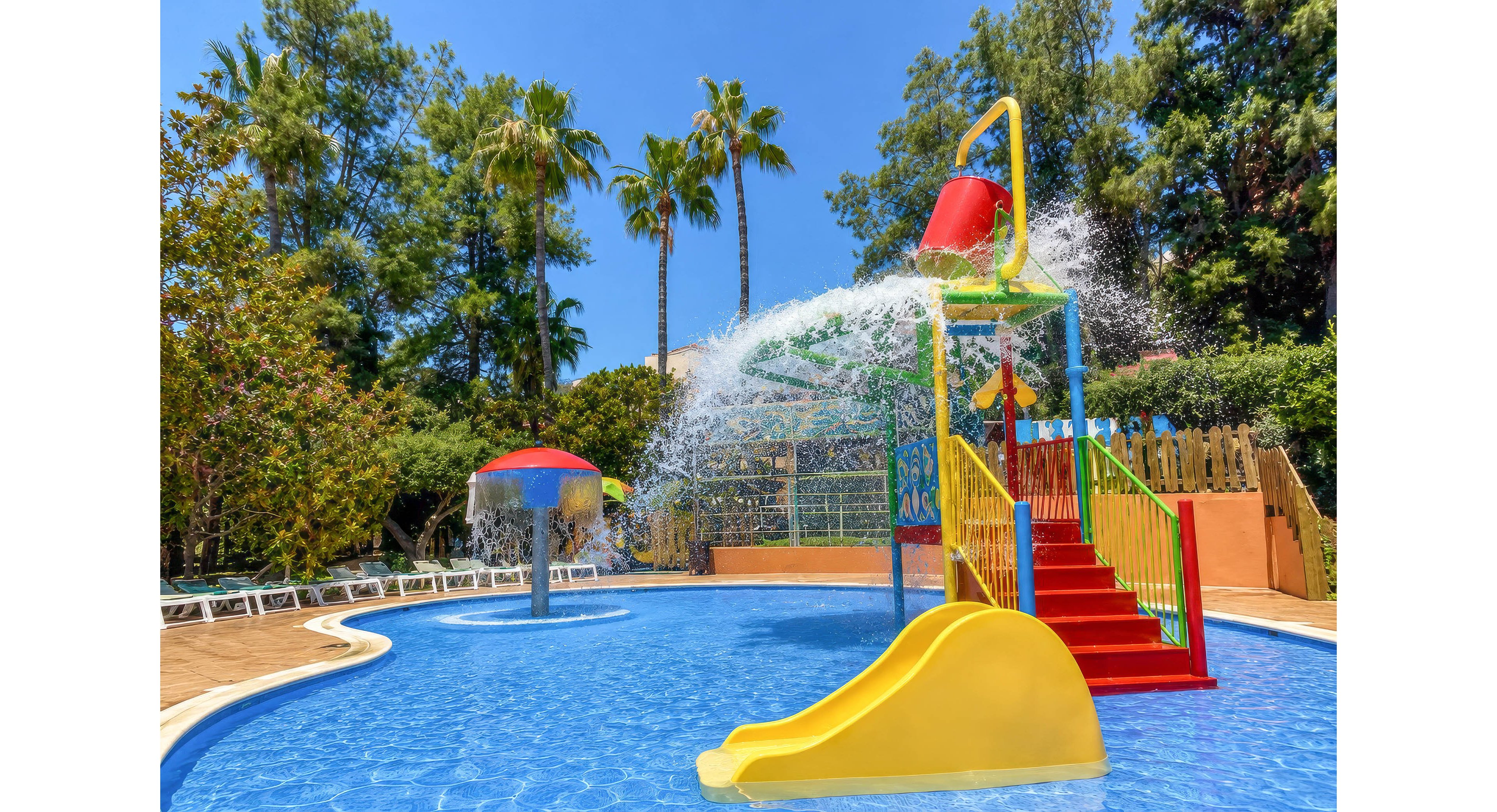 A vibrant children’s water play area features a yellow slide and red stairs leading up to a large tipping bucket. Water splashes down into a shallow blue pool surrounded by trees and greenery. Additional play structures and fountains are visible in the background, along with sun loungers along the poolside. The scene is bright and sunny, creating a fun and cheerful atmosphere.