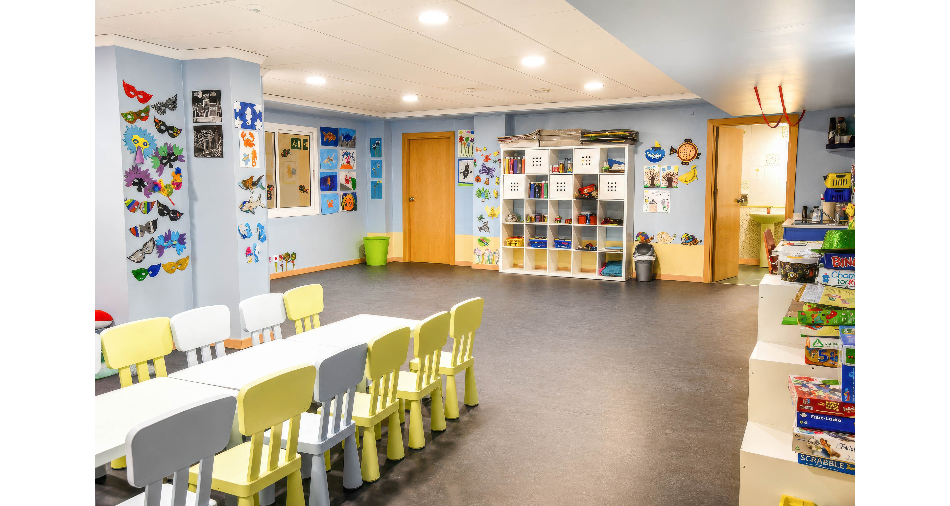 Colorful classroom interior features a long white table surrounded by small pastel chairs. The walls are decorated with children’s artwork, masks, and drawings, creating a playful learning environment. Open cubby shelves hold storage bins and toys, while additional games and supplies are stacked on a side unit. The room is well-lit with ceiling lights and has doors leading to adjacent spaces, suggesting a daycare or preschool setting.