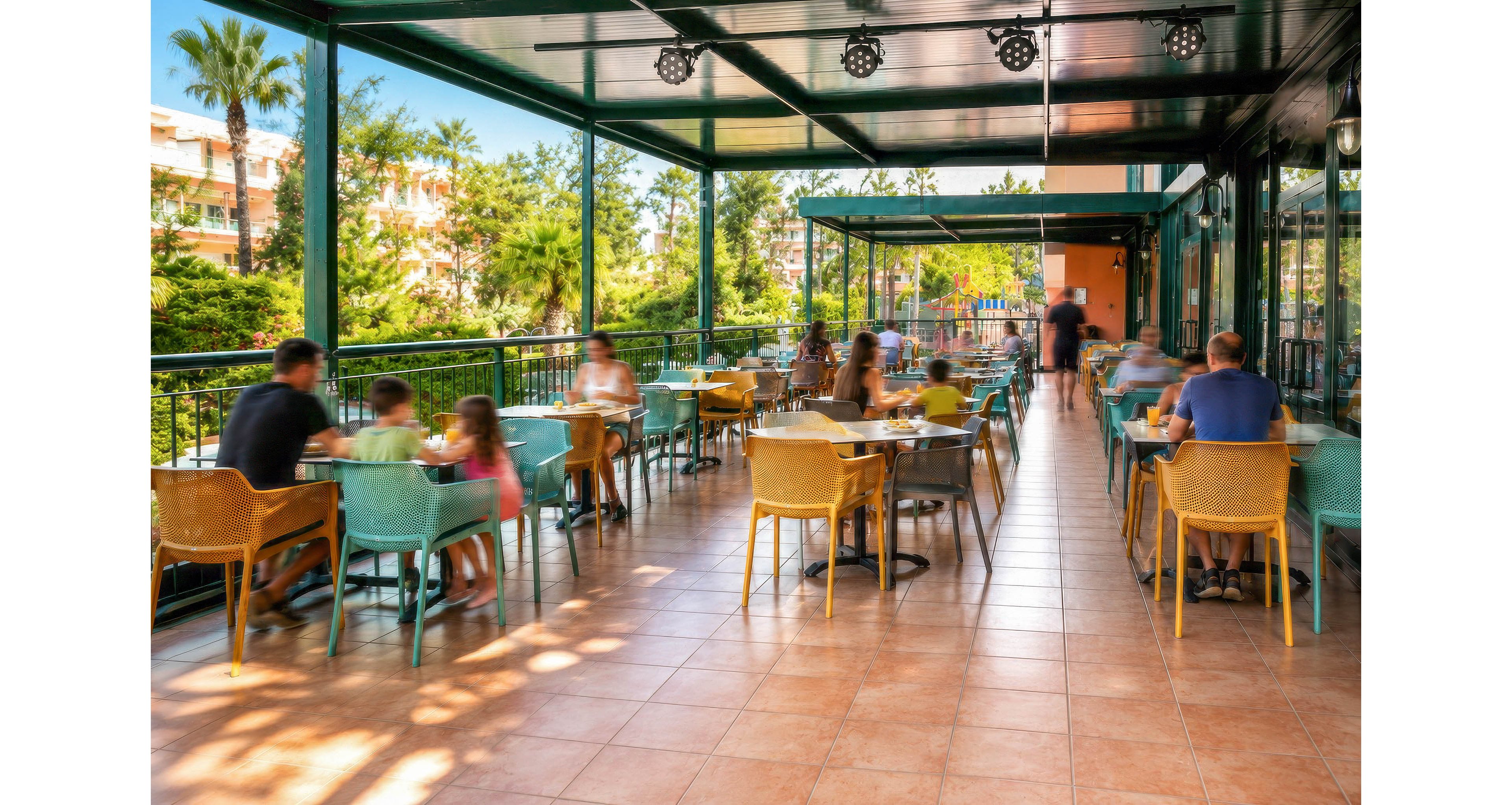 An open-air terrace café features colorful chairs and tables arranged along a tiled floor. Adults and children sit and talk while enjoying food and drinks in a relaxed setting. The space is covered by a modern green metal roof with lighting fixtures and bordered by railings overlooking lush greenery. Warm sunlight and vibrant colors create a casual, inviting atmosphere suitable for socializing and leisure.