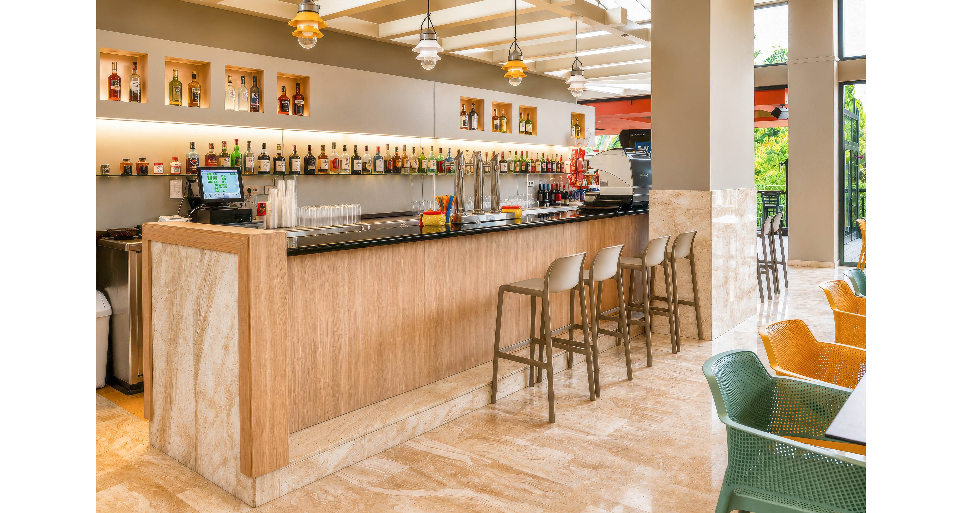 A contemporary bar counter with a polished black top and light wood paneling stands in a bright indoor space. Bottles of liquor are neatly arranged on backlit shelves behind the bar, with a point-of-sale screen and drink dispensers on the counter. Several high stools line the bar, while colorful mesh chairs and tables occupy the foreground seating area. Large windows and open walls allow natural light to fill the room, creating an inviting atmosphere.