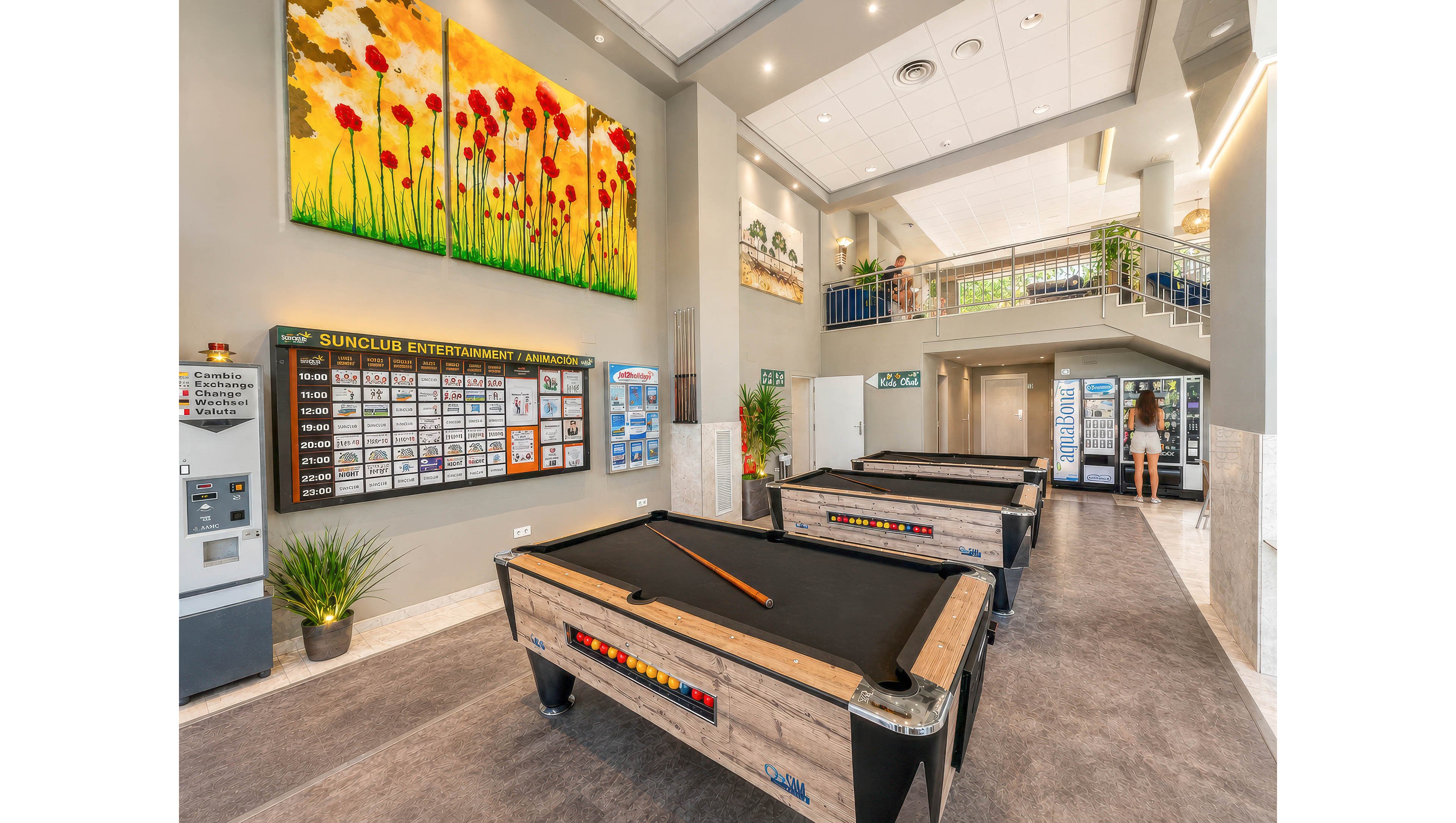 Spacious indoor entertainment room featuring several wooden pool tables as the main focus. A large colorful triptych painting with red flowers hangs above a detailed SUNCLUB entertainment timetable board listing activities and times. The setting includes bright lighting, potted plants, and a mezzanine level with railings and additional decor. A person is visible near the entrance area, adding a casual leisure atmosphere to the scene.