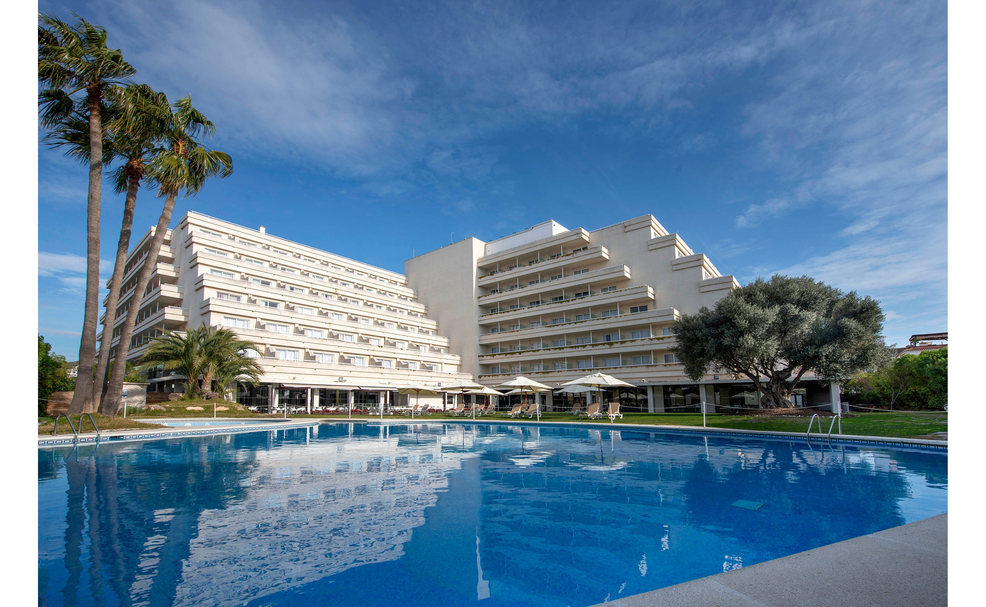 A large modern resort hotel stands beside a clear blue outdoor swimming pool. The multi-story buildings feature wide balconies and a clean, geometric design. Palm trees and other greenery surround the pool area, with sunshades and seating visible along the edge. The scene is captured under a bright blue sky with scattered clouds, creating a calm vacation atmosphere.