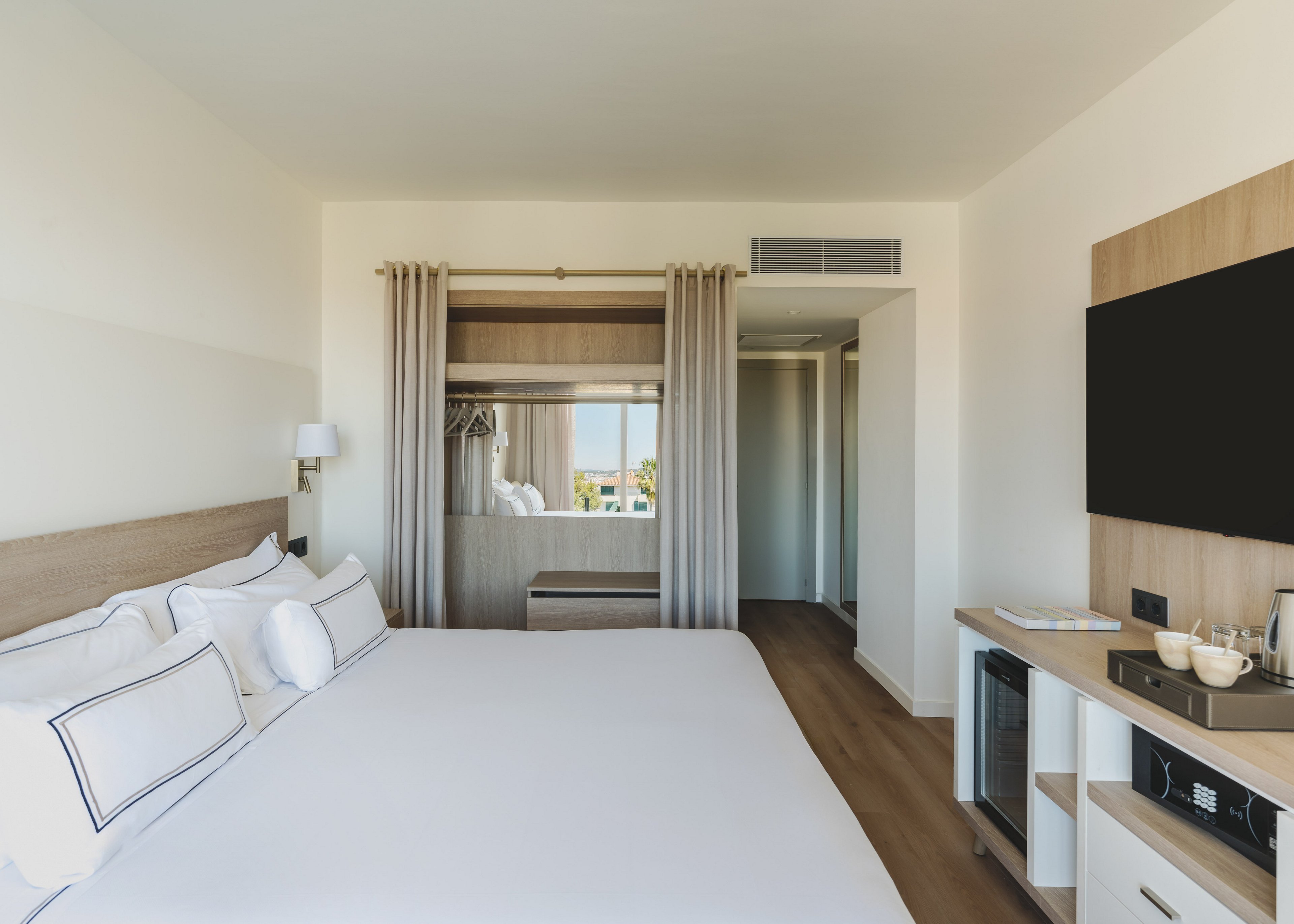 A bright, modern hotel room features a large neatly made bed with white linens and accent pillows. Opposite the bed, a flat-screen television is mounted above a light wood console with a minibar and coffee setup. Neutral curtains frame a window and an open wardrobe area with hangers and a bench. The room has a minimalist design with clean lines, soft lighting, and a calm, neutral color palette.