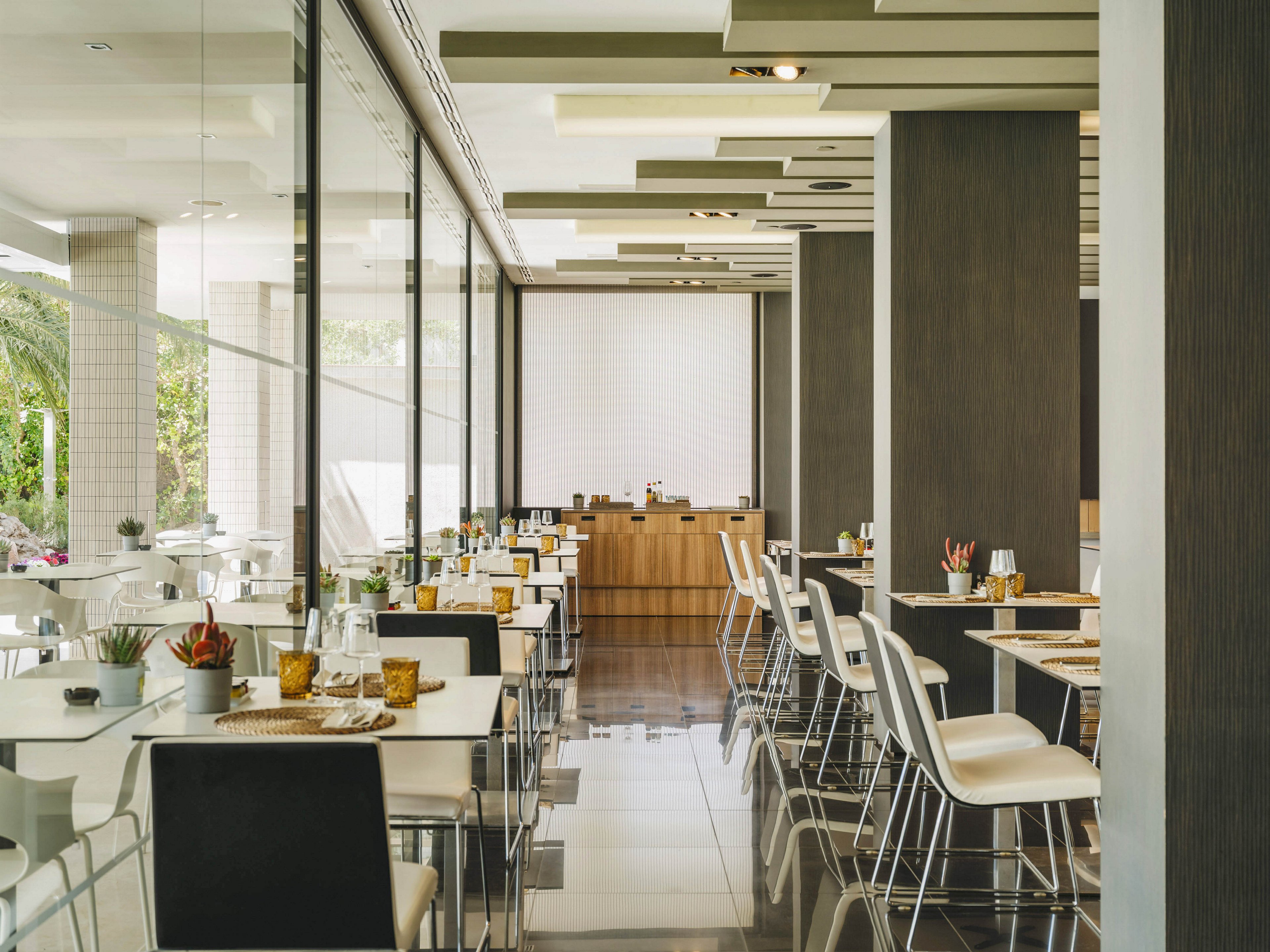 A contemporary restaurant dining area features neatly arranged white tables and chairs. Large floor-to-ceiling glass windows line one side, allowing natural light to brighten the space and reveal greenery outside. The polished floor reflects the ceiling panels and minimalist decor, creating a clean and sophisticated atmosphere. Tabletop details such as small plants, candles, and place settings add warmth to the modern interior.