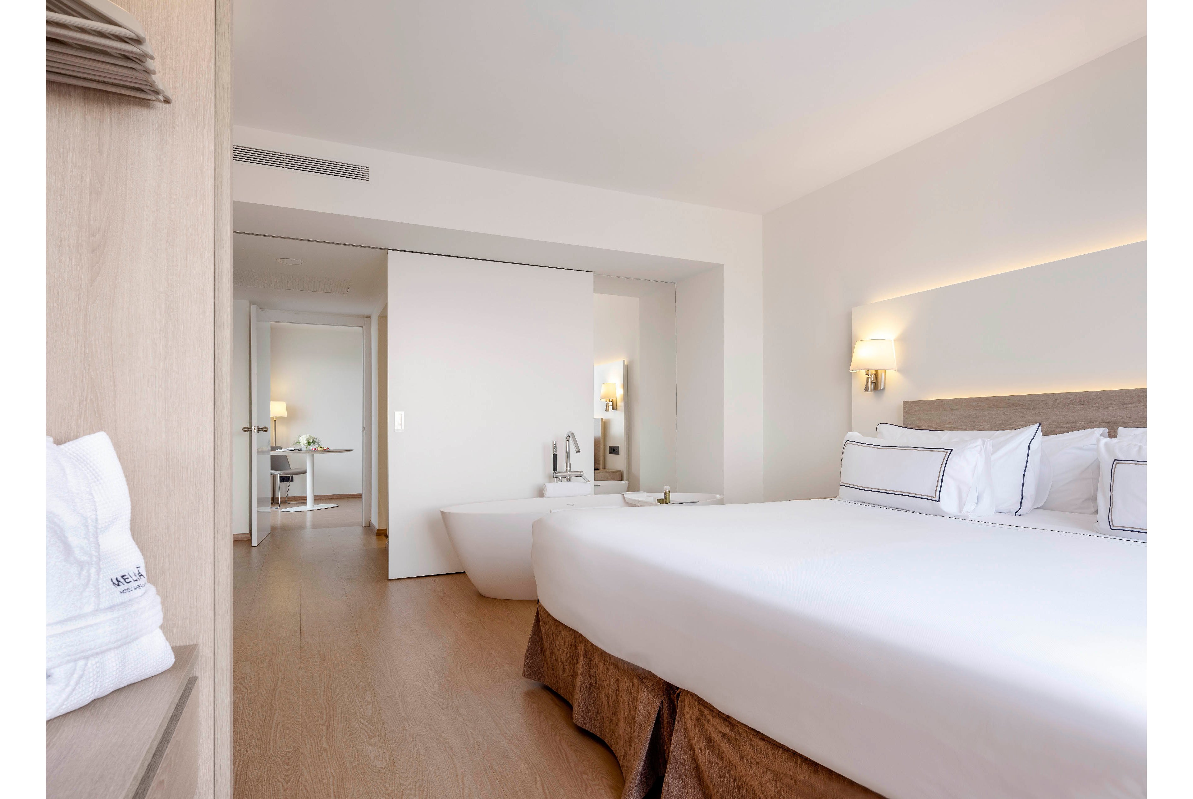 A bright, modern hotel bedroom features a large neatly made bed with white linens and neutral-toned furnishings. A freestanding bathtub is positioned near a partially open sliding door leading to an adjacent room. Soft lighting from wall sconces and a bedside lamp creates a calm, inviting atmosphere. The minimalist decor and light wood flooring emphasize a clean, contemporary design.
