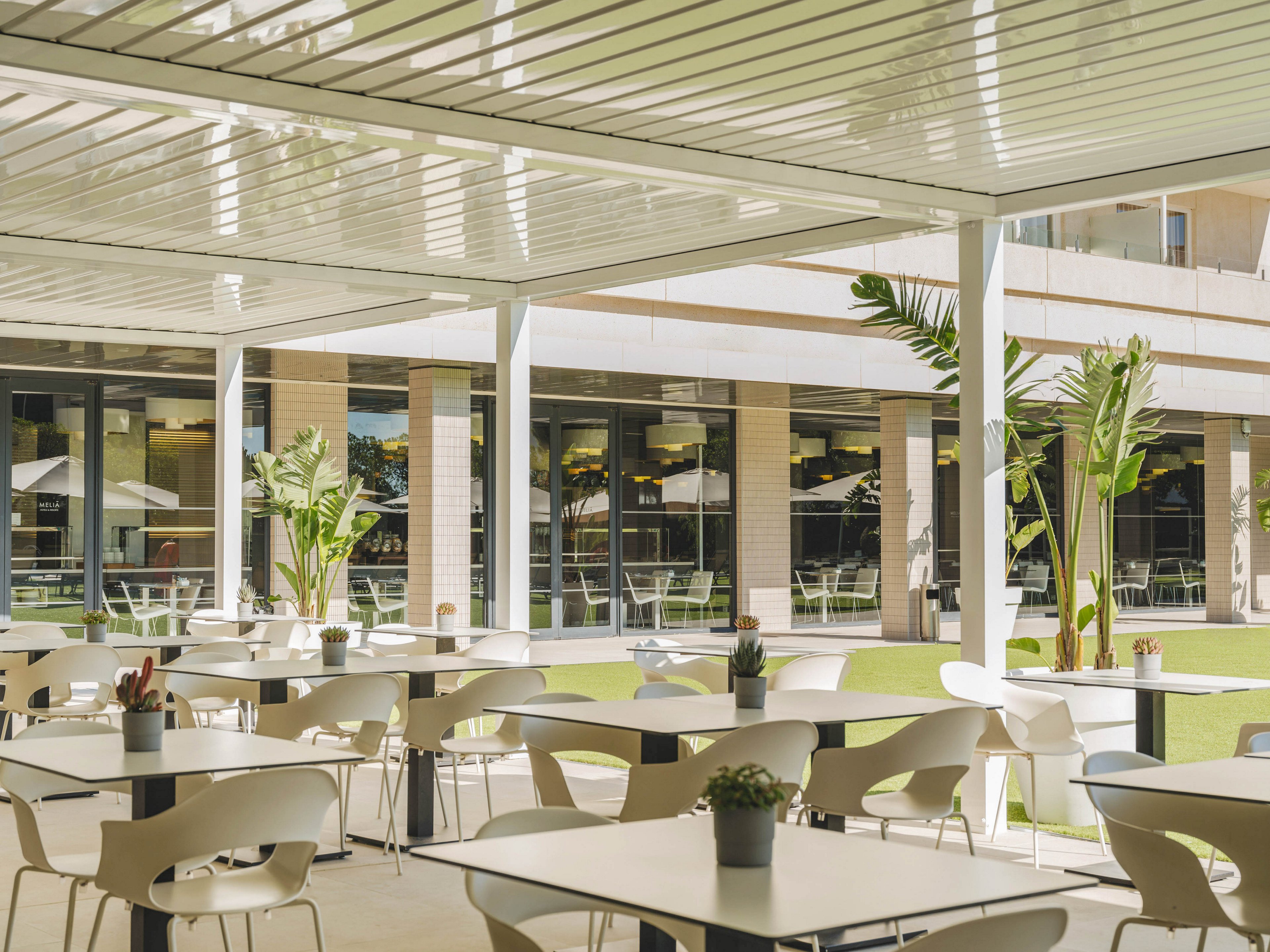 A contemporary outdoor cafe terrace features rows of white tables and chairs under a sleek pergola roof. Potted plants decorate the tables while larger tropical plants line the edge of the seating area. In the background, large glass windows reveal an indoor space and additional seating on a green lawn. The scene is bright and airy, suggesting a relaxed and welcoming atmosphere.
