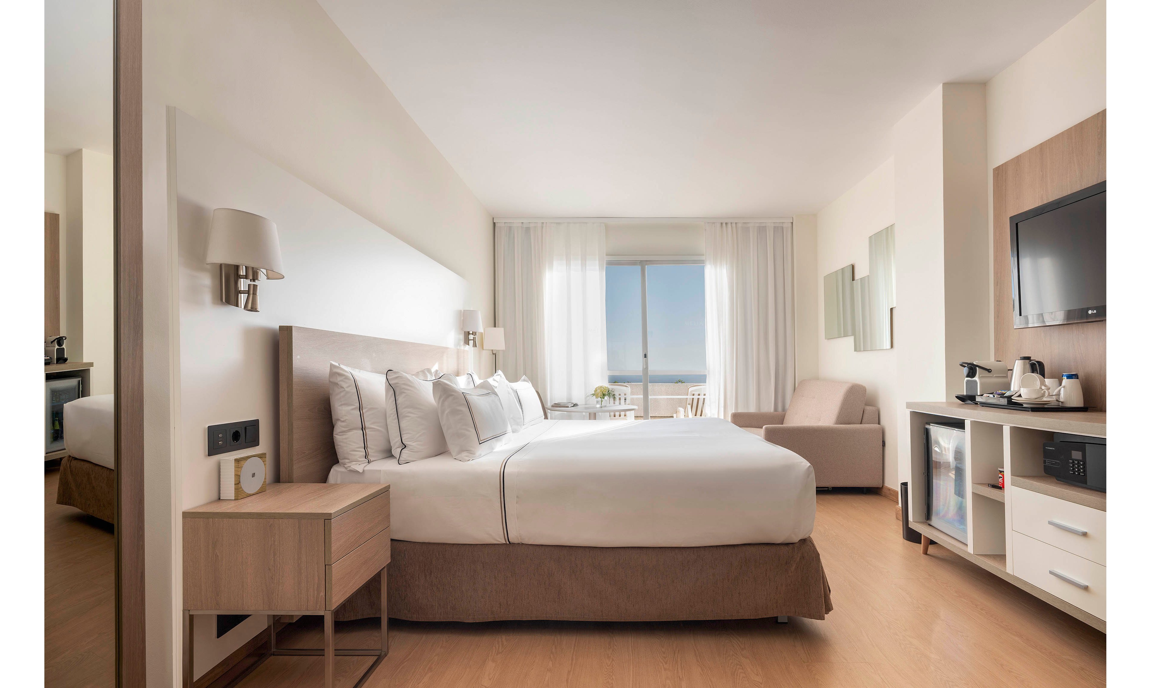 A contemporary hotel room features a large neatly made bed with multiple pillows and neutral bedding. The space includes a bedside table, wall-mounted lamps, and a compact entertainment unit with a flat-screen TV, minibar, and coffee station. A soft armchair sits near a large window framed by sheer white curtains, revealing a distant view of the sea and sky. The room’s light color palette and clean lines create a calm, inviting atmosphere suitable for relaxation or business travel.