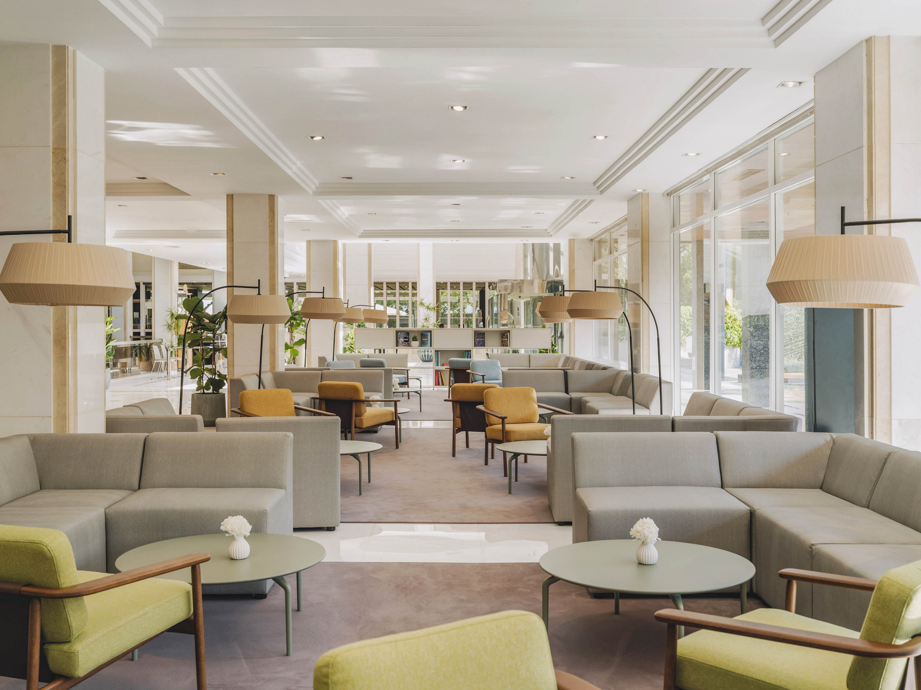 A spacious, modern hotel lobby features light gray sectional sofas and pastel green armchairs arranged around low circular tables. Large floor-to-ceiling windows on one side allow natural light to flood the space, highlighting the neutral tones and clean lines. Oversized pendant lamps and floor lamps provide additional soft lighting, creating a welcoming atmosphere. Small white flower arrangements on the tables add a subtle decorative touch to the minimalist interior.