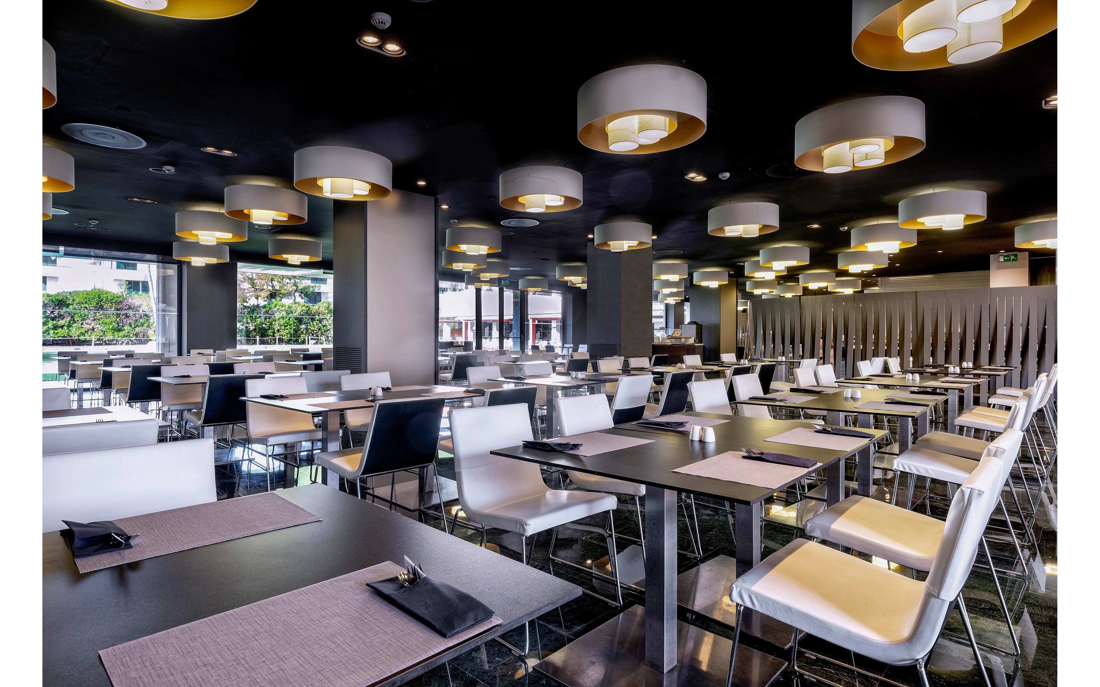 A spacious, modern restaurant dining area is shown with rows of neatly arranged tables and chairs. The room features sleek black tables, white chairs, and placemats set in preparation for guests. Large circular ceiling lights create a warm, inviting glow against the dark ceiling. Floor-to-ceiling windows in the background reveal an urban outdoor setting, adding natural light to the interior.