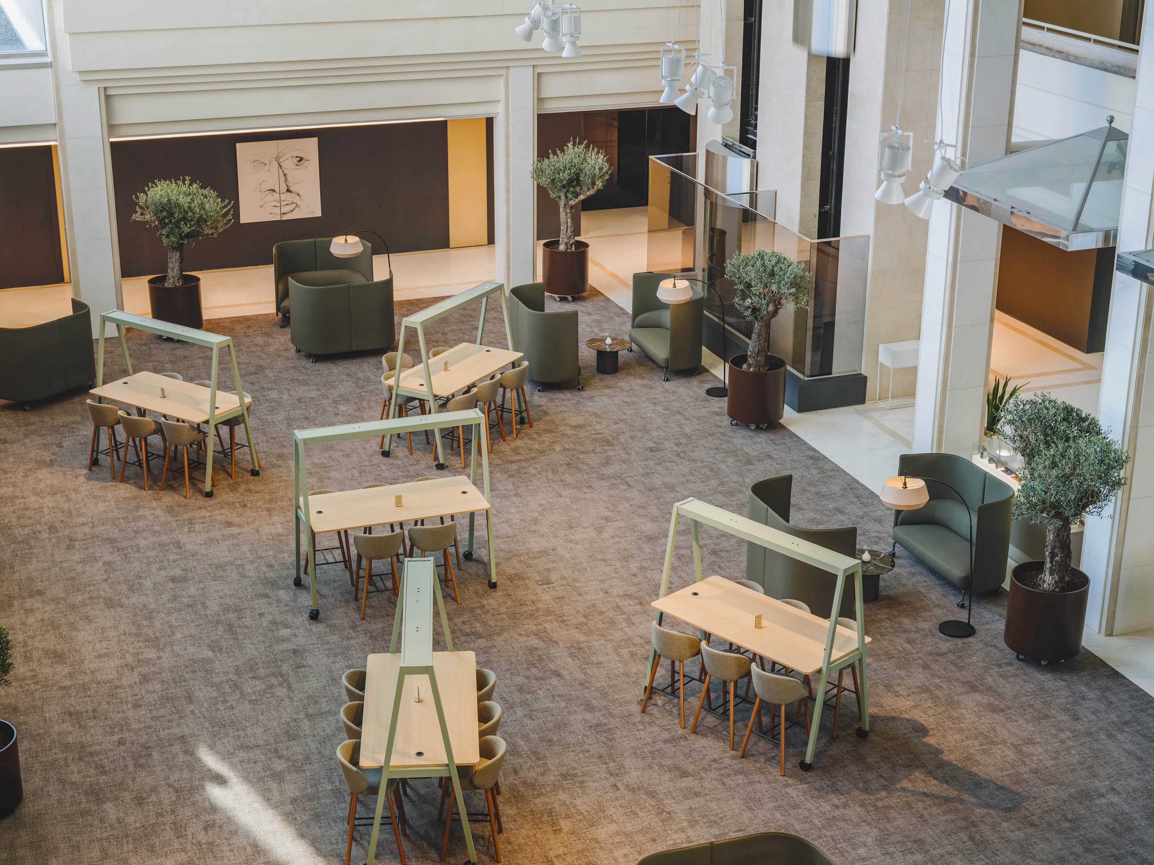 A spacious modern lobby features several light-colored communal tables arranged in an open layout. Soft armchairs and small round side tables line the perimeter, creating relaxed seating zones. Large potted trees add greenery and contrast against the neutral carpet and walls. Natural light filters in from tall windows, giving the space an airy, contemporary feel.