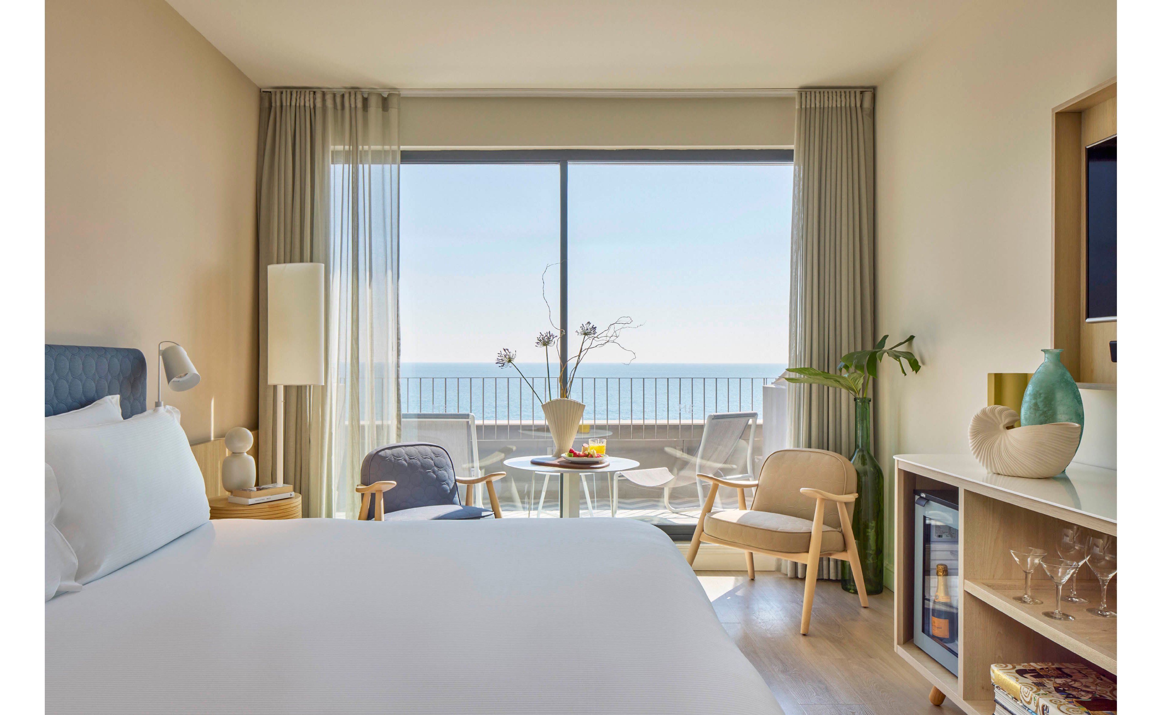 A modern hotel room features a neatly made bed facing a large window with a clear ocean view. Neutral-toned furniture, including armchairs and a small round table, create a cozy seating area by the balcony. Soft natural light filters through sheer curtains, highlighting decorative vases, plants, and a compact minibar. The overall atmosphere feels calm, airy, and inviting, ideal for a relaxing seaside stay.