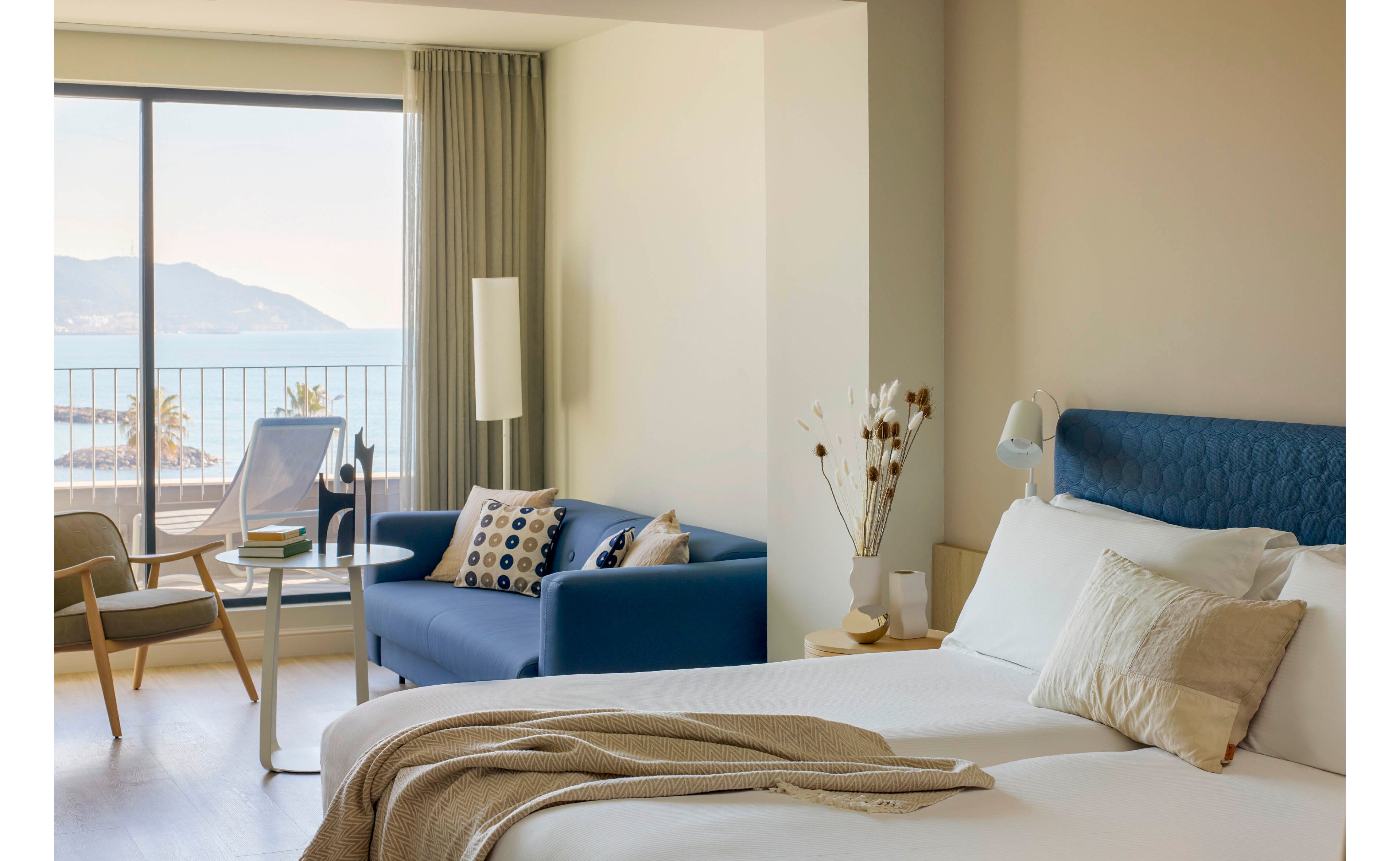A modern hotel room features a neatly made bed with neutral bedding and a blue upholstered headboard. A blue sofa with patterned cushions and a small coffee table sit near a large window. The window opens to a balcony overlooking the sea and distant mountains, allowing natural light to fill the space. Soft beige walls, light wood furniture, and minimalist decor create a calm, contemporary atmosphere.