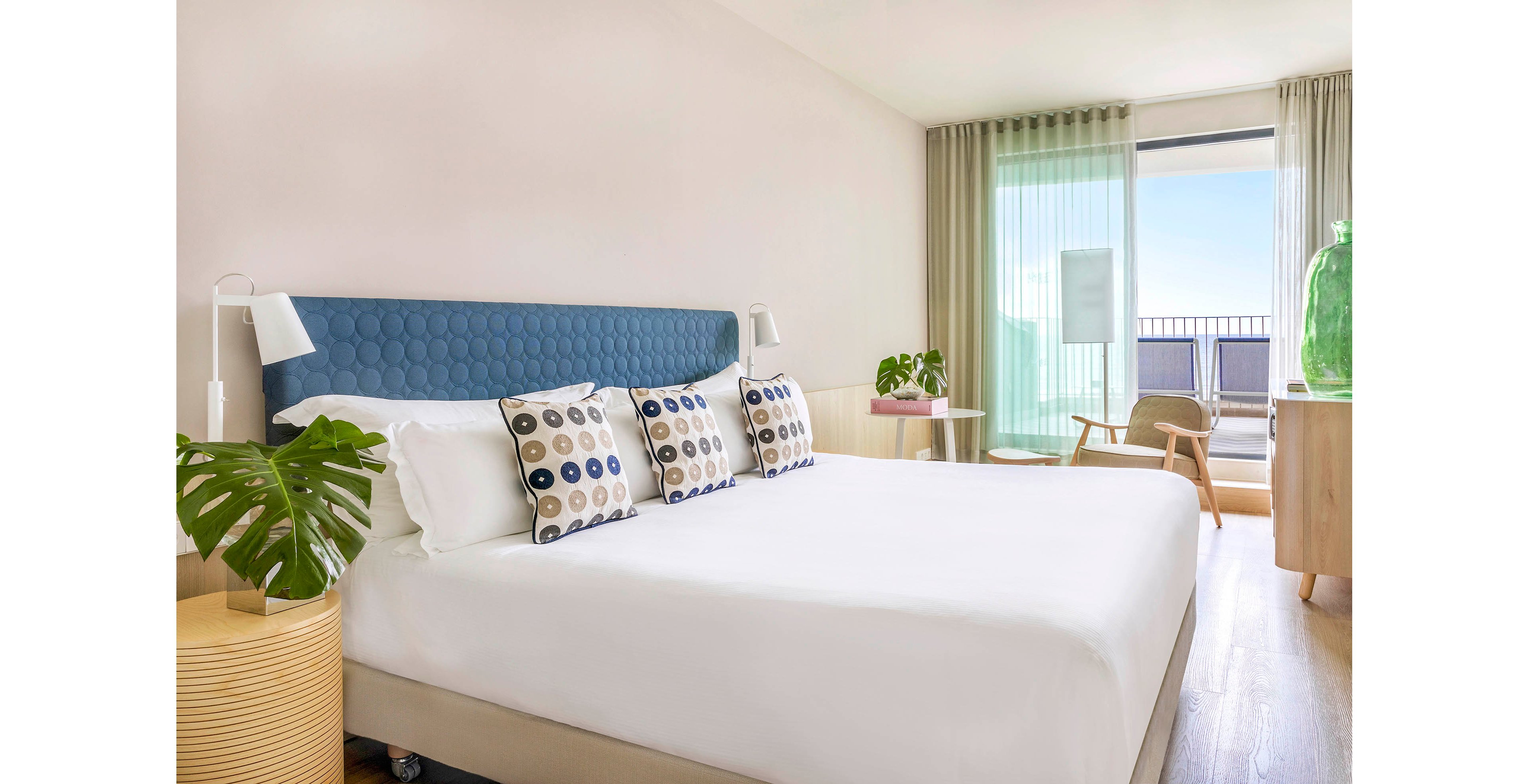 A contemporary hotel bedroom features a large neatly made bed with a blue upholstered headboard and patterned pillows. The room is decorated in light neutral tones with wooden flooring and minimalist furniture, including a bedside table and a lounge chair. Large windows with sheer curtains open to a balcony, allowing natural light to fill the space and reveal a distant view. Green plants and a glass decorative piece add fresh accents to the clean, modern interior.