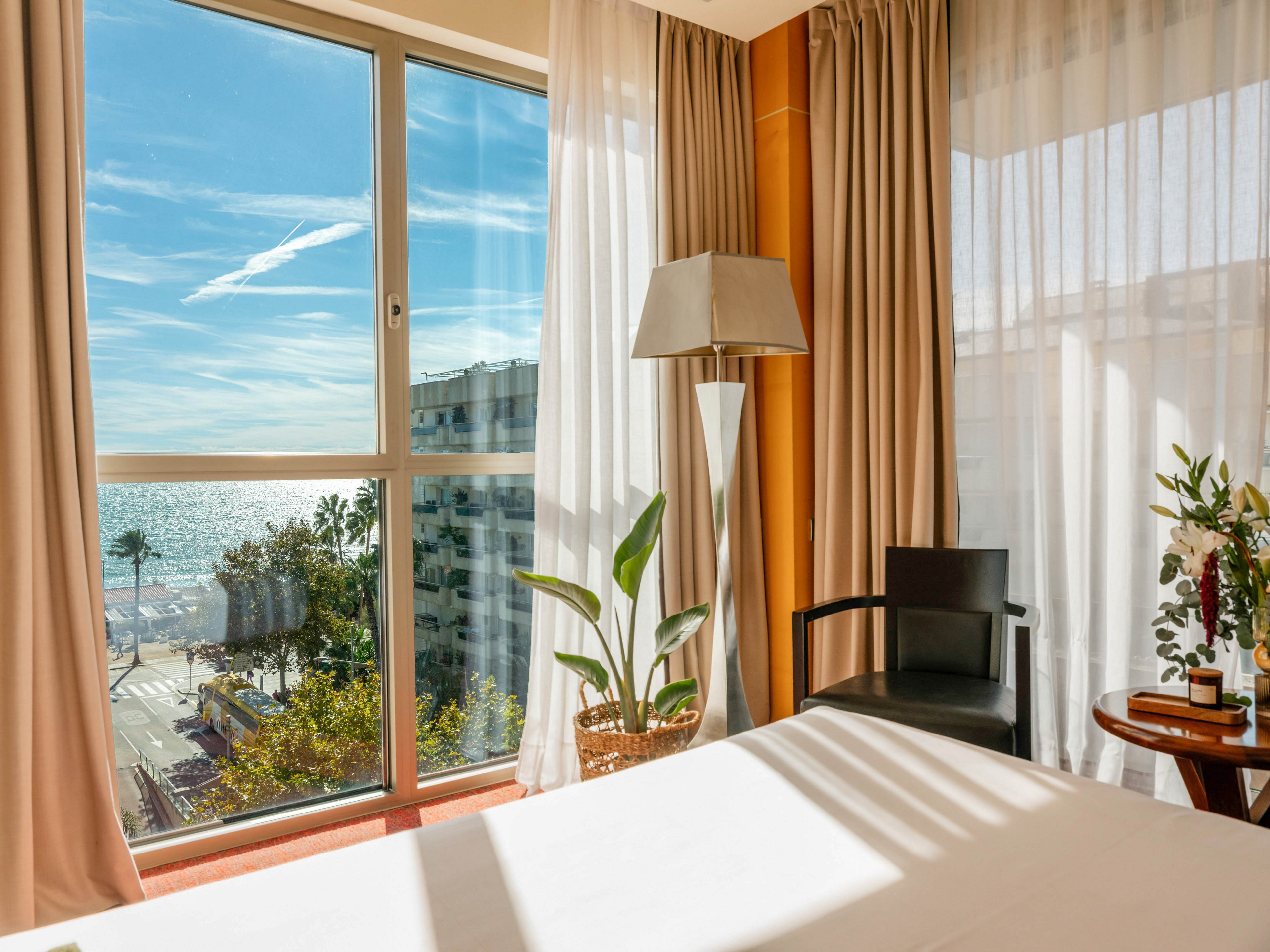 A bright hotel room features a large bed positioned near a floor-to-ceiling window. Sunlight streams through sheer curtains, revealing a coastal city street and ocean in the distance. A modern floor lamp, black chair, and potted plant add contemporary touches to the interior. The warm color palette and clear blue sky outside create a relaxed, vacation-like atmosphere.