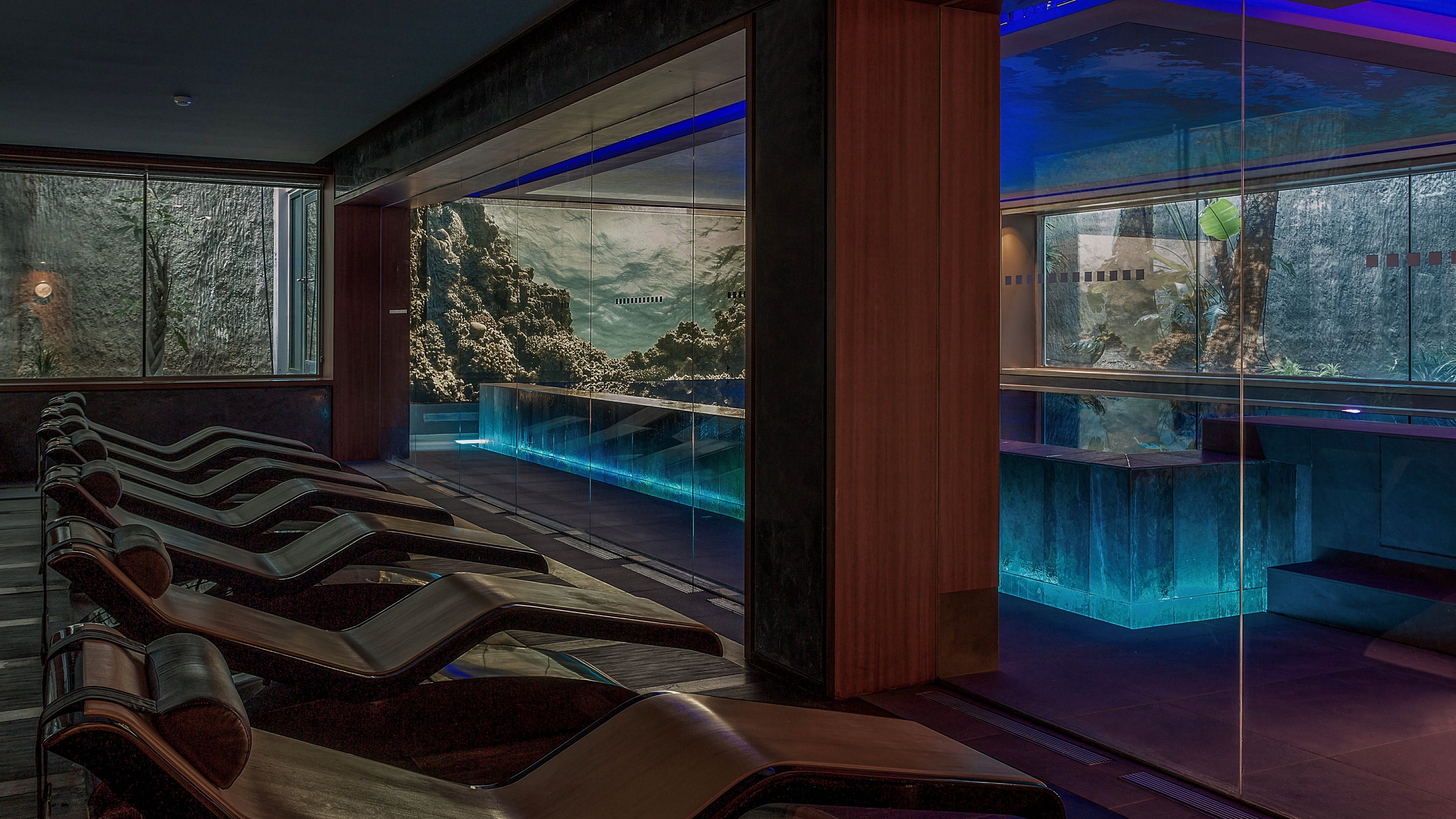A contemporary spa lounge features a row of reclining loungers facing large glass windows. Beyond the windows, a rocky outdoor landscape with trees is visible, creating a natural backdrop. Inside, long rectangular pools and counters are lit with blue underwater lighting, casting a cool glow across the dark interior. The overall atmosphere is tranquil and luxurious, emphasizing relaxation and modern design.