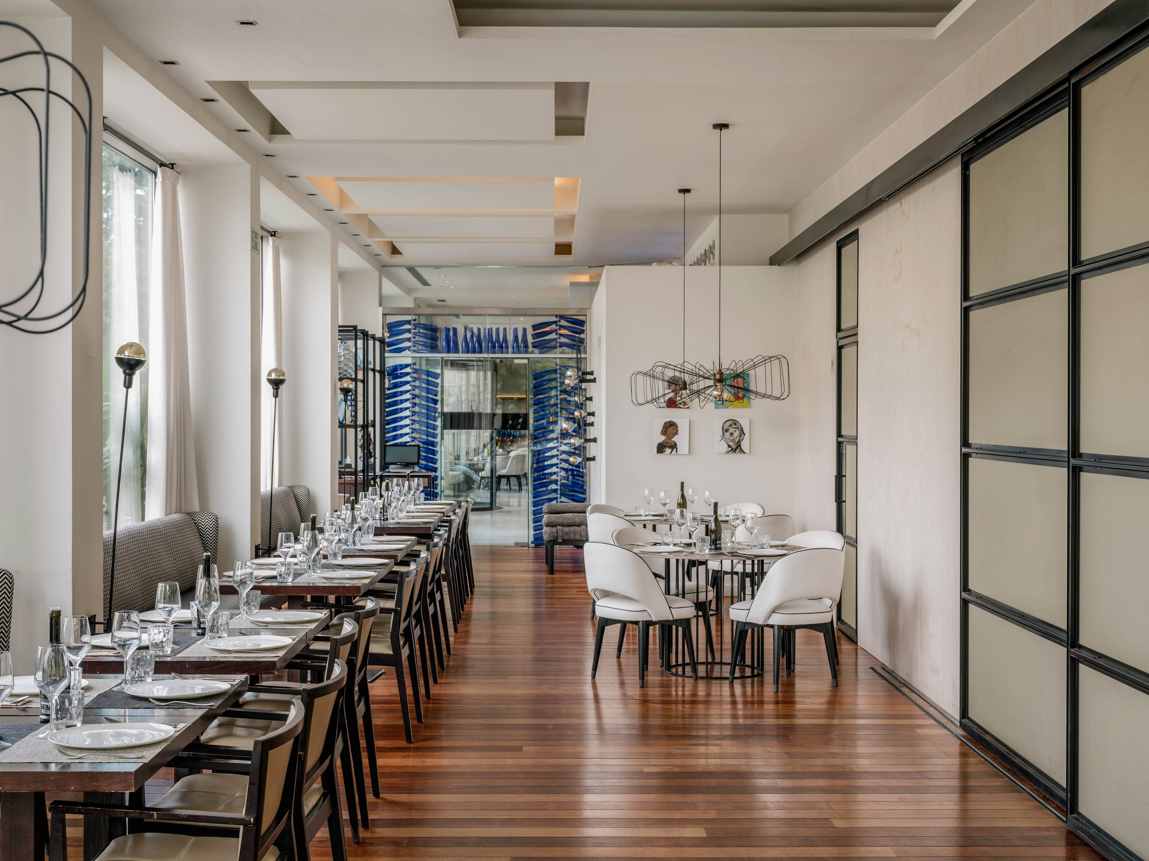 Contemporary restaurant dining room with neatly arranged tables and chairs stands empty, ready for guests. Large windows on the left bring in natural light, illuminating the polished wooden floor and neutral-toned walls. A round table with white chairs and decorative light fixtures creates a focal point in the center. The overall atmosphere is clean, sophisticated, and inviting, suitable for fine dining or special occasions.
