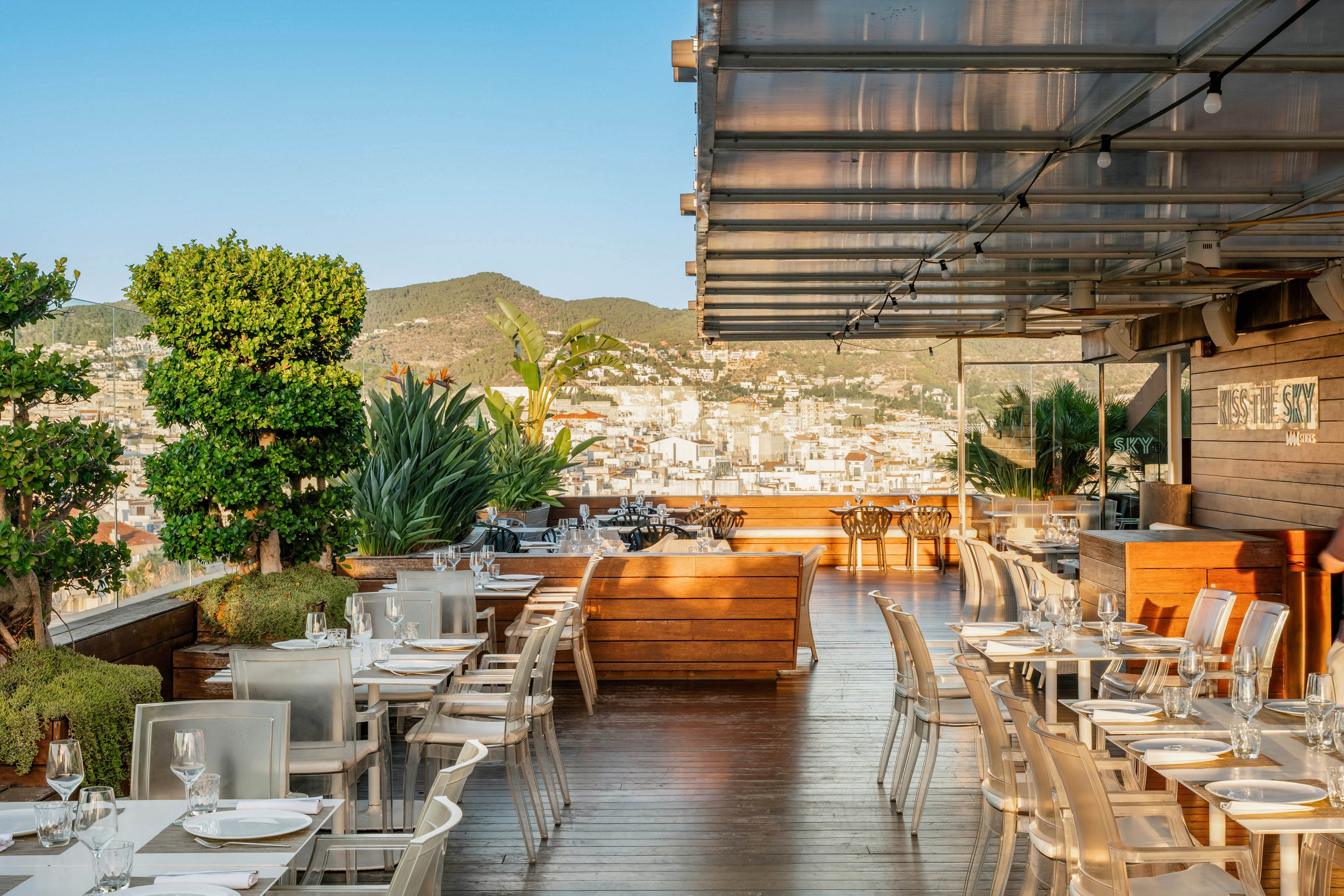 An elegant rooftop restaurant terrace is set with neatly arranged tables and clear chairs. Wooden planters with lush green plants line the edge, framing a panoramic view of the city and surrounding hills. Glassware and table settings glint in the warm sunlight under a partially covered pergola. The atmosphere feels relaxed and upscale, ideal for outdoor dining with scenic urban views.