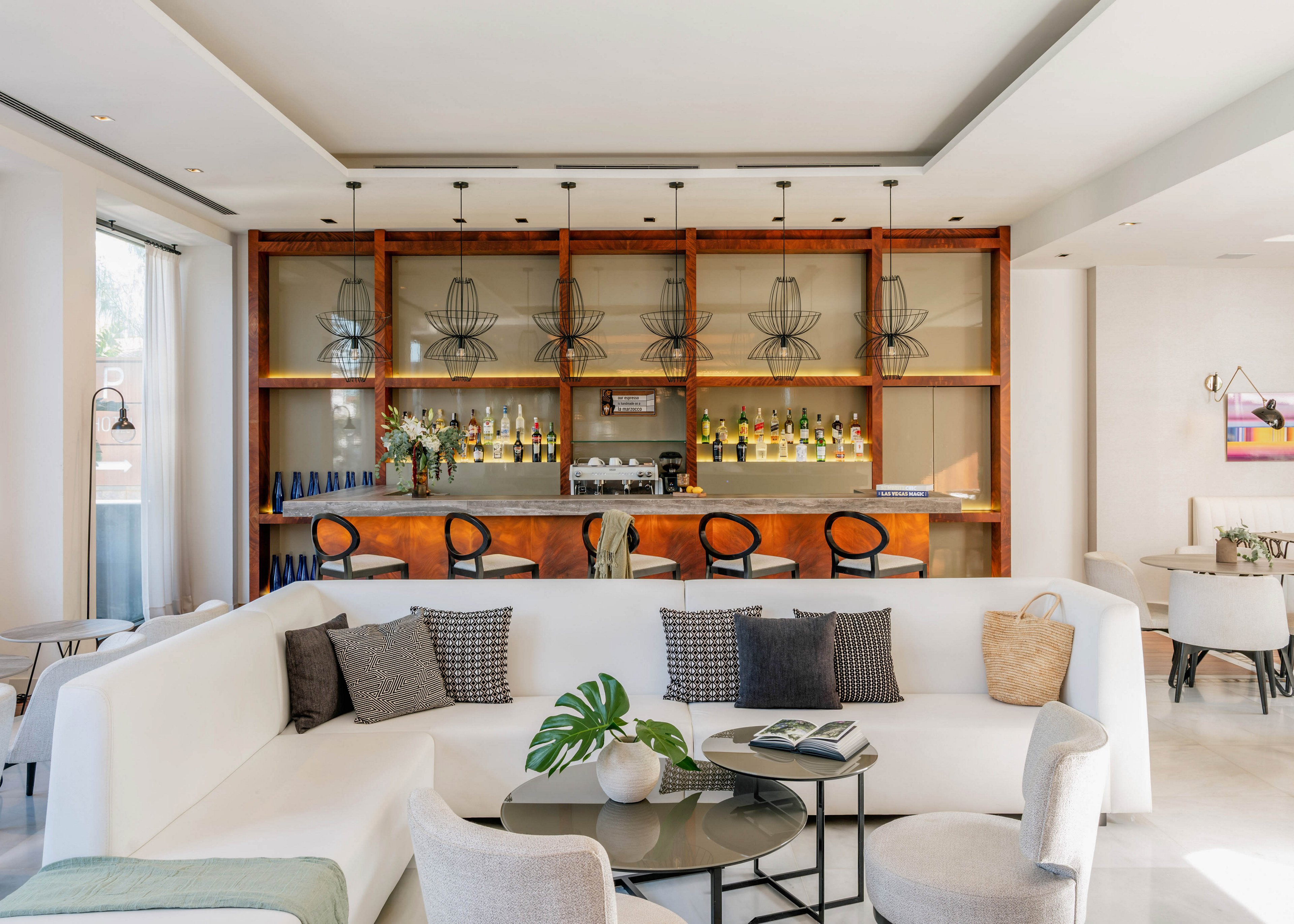 A contemporary hotel lounge features a sleek bar lined with bottles and glassware in the background. In the foreground, a white sectional sofa and light upholstered chairs surround a low coffee table with books and a green plant. Warm wood shelving, pendant lights, and neutral decor create an inviting, minimalist atmosphere. The space is bright and airy, suggesting a relaxed social or waiting area.