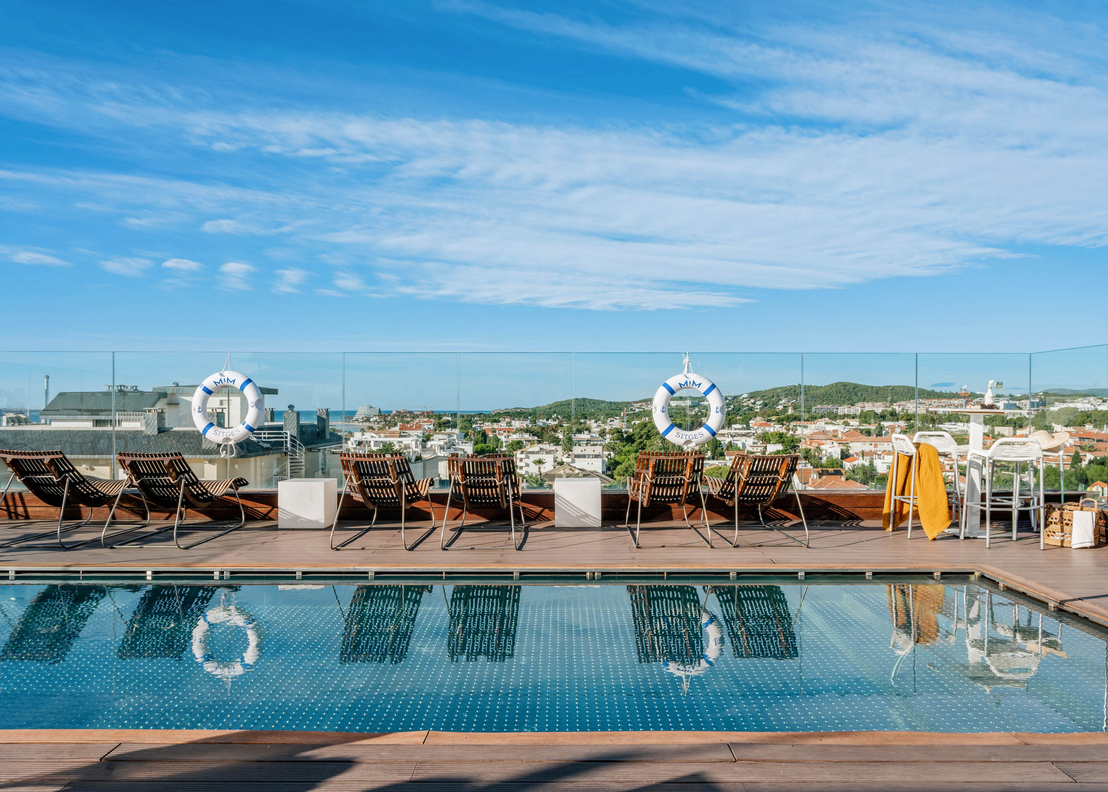 A rooftop swimming pool reflects the blue sky and scattered clouds on a clear day. Wooden lounge chairs with cushions are arranged along the poolside, some with towels draped over them. Glass safety railings surround the terrace, offering an open view of the distant cityscape and green hills. Two life preservers hang on the railing, adding a nautical accent to the modern outdoor setting.