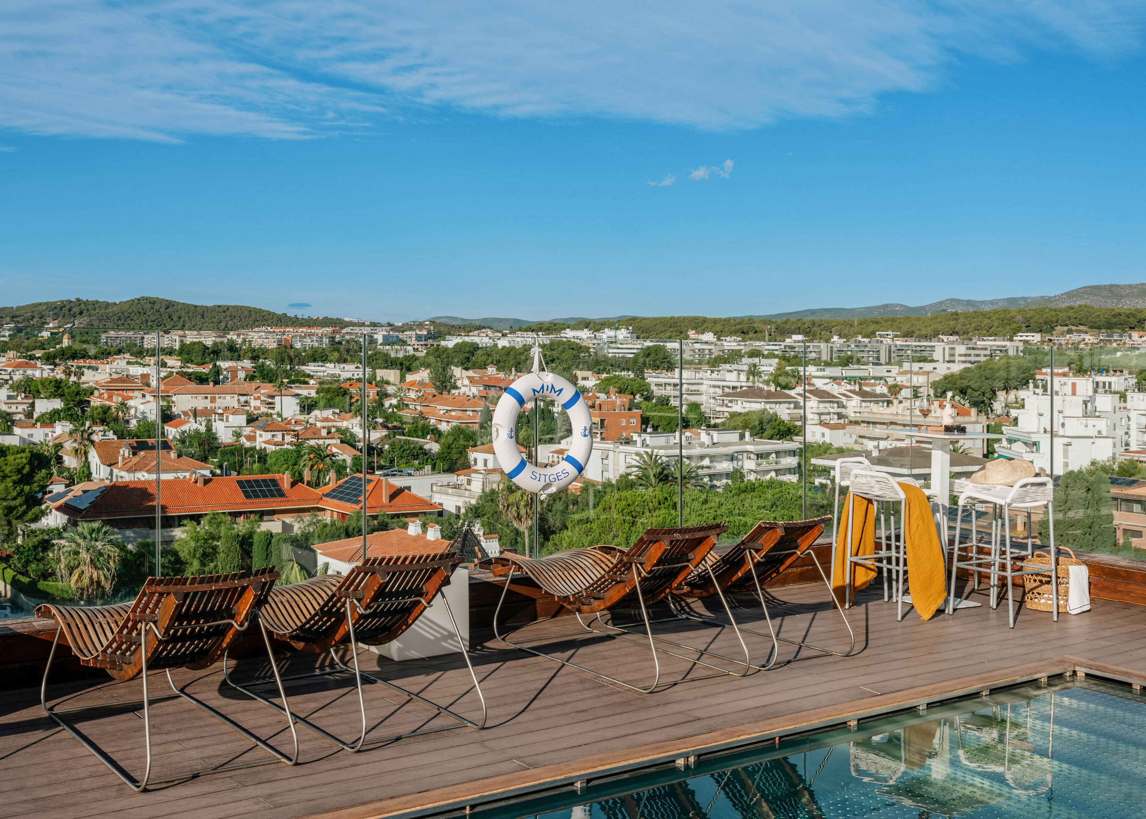 A modern rooftop pool terrace features a row of wooden lounge chairs facing a glass railing. Beyond the deck, a sprawling cityscape with white buildings, red roofs, and green trees stretches into the distance under a bright blue sky. A white lifebuoy hangs on the railing, and a small bar area with stools and towels adds to the relaxed atmosphere. The scene conveys a sunny, leisurely urban escape with panoramic views.