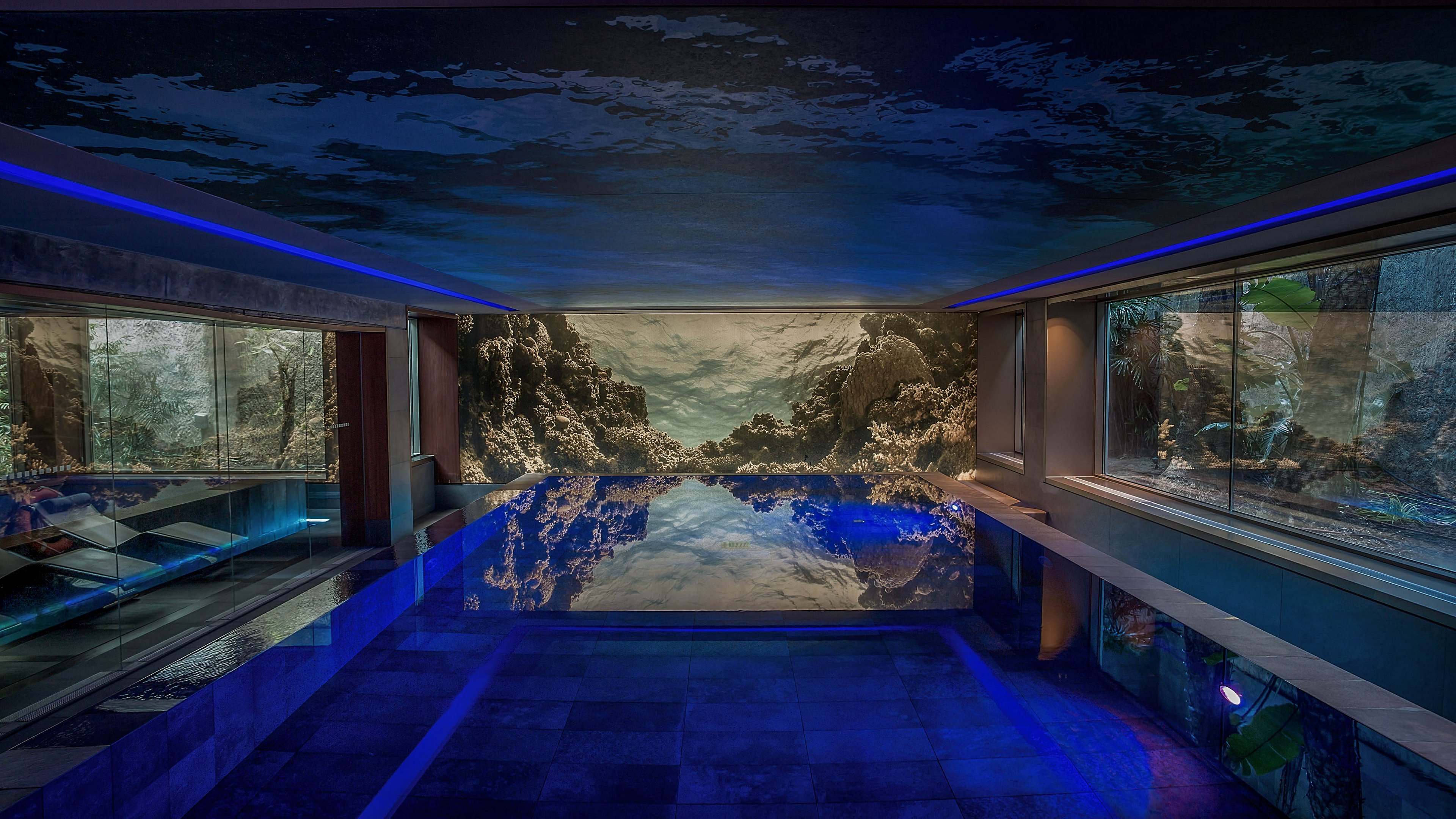 A luxurious indoor swimming pool is illuminated with dramatic blue accent lighting. Large panoramic wall murals depict an underwater or rocky seascape, reflected on the still surface of the water. Floor-to-ceiling windows on the right side reveal additional aquatic-themed visuals or aquariums. The overall atmosphere is modern, immersive, and tranquil, emphasizing relaxation and high-end design.