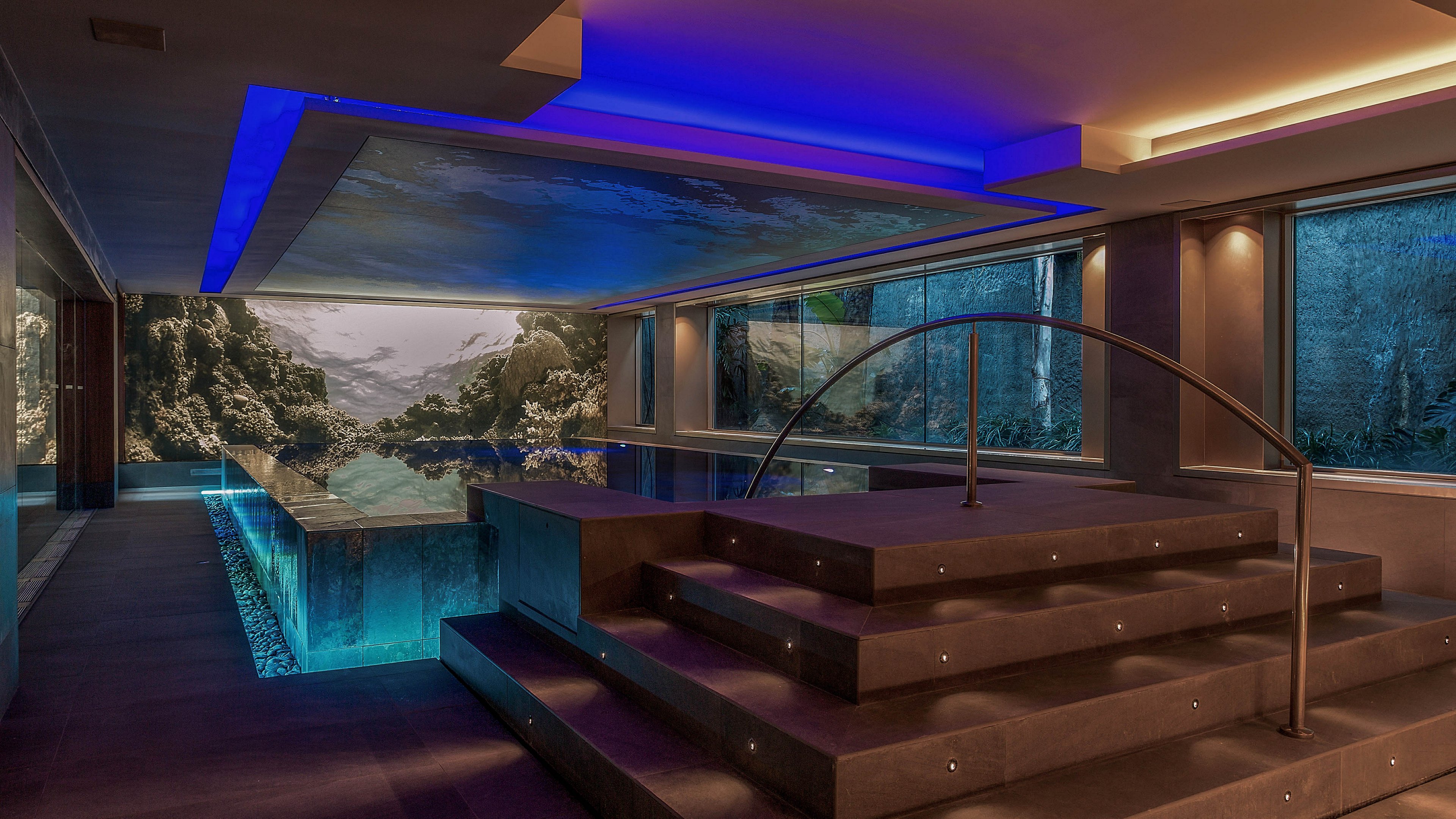 A luxurious indoor swimming pool is set within a contemporary spa-like interior. Wide steps with integrated lights lead down to the water, framed by sleek metal handrails. Large floor-to-ceiling windows reveal a rocky natural landscape outside, enhancing the tranquil atmosphere. Blue and warm-toned ceiling lights create a dramatic contrast and reflect softly on the pool surface.
