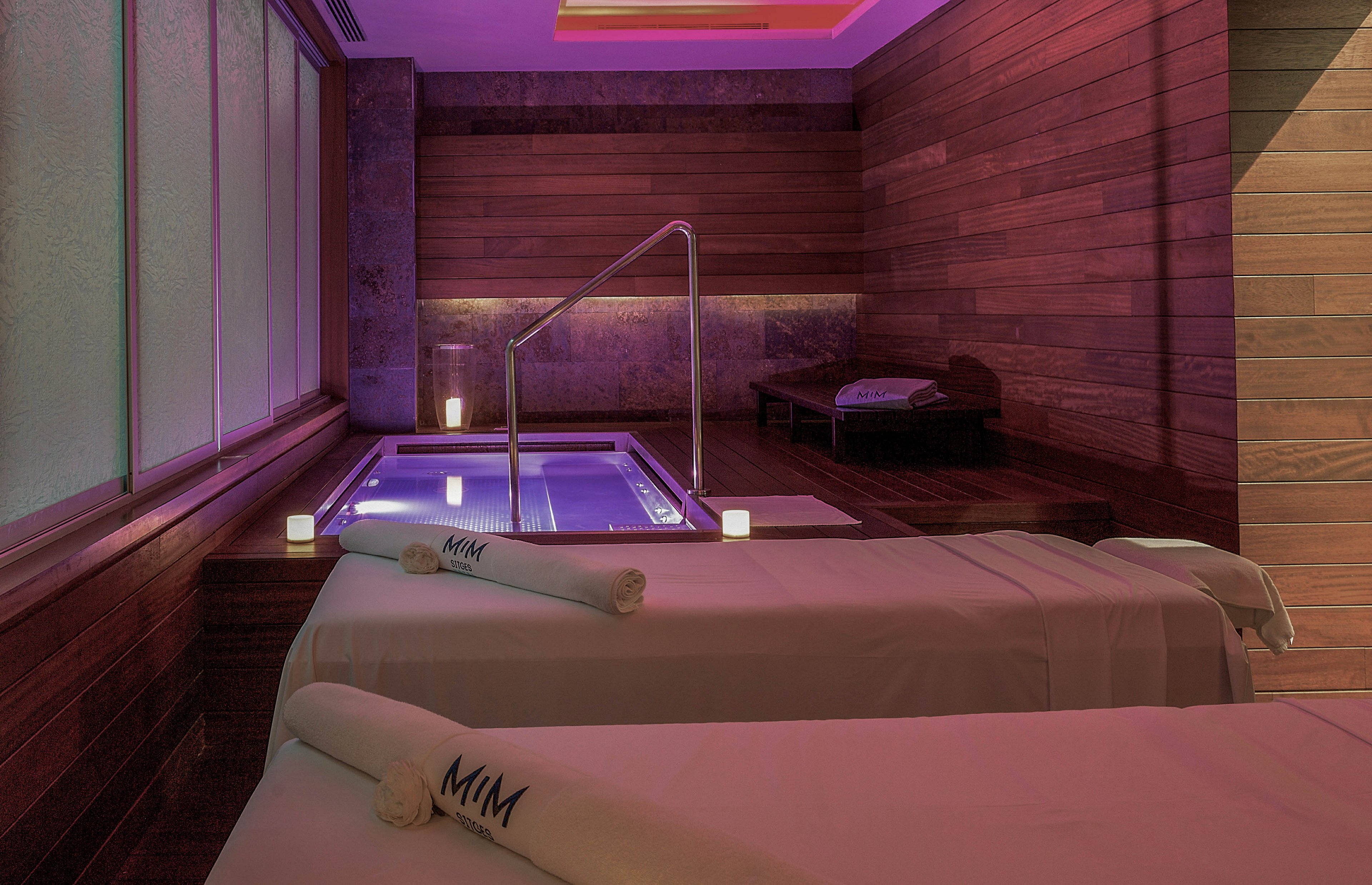 A serene spa room features two massage tables in the foreground, each topped with neatly rolled towels marked with the letters 'MIM'. In the background, a small illuminated jacuzzi pool with metal handrails is set into a wooden floor. Warm wooden wall panels and soft purple ambient lighting create a relaxing, upscale atmosphere. The overall scene suggests a tranquil wellness retreat prepared for guests.