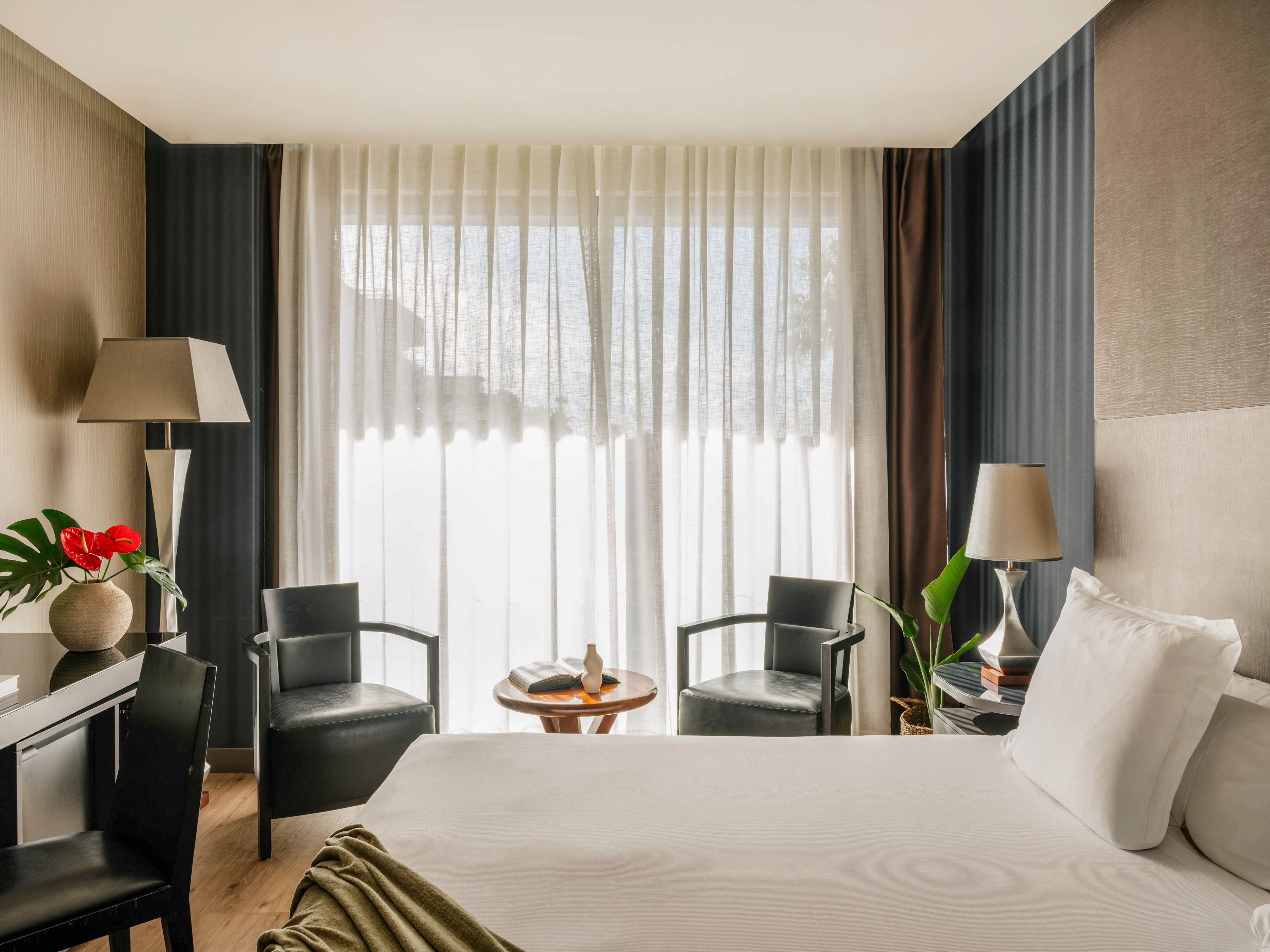 A contemporary hotel room features a neatly made bed facing a large window with sheer curtains. Two black armchairs and a small round table create a cozy seating area by the natural light. A desk with a lamp, vase, and red flowers sits to the side, while another floor lamp and green plant add warmth to the space. Neutral tones with dark accents give the room a calm, modern atmosphere.