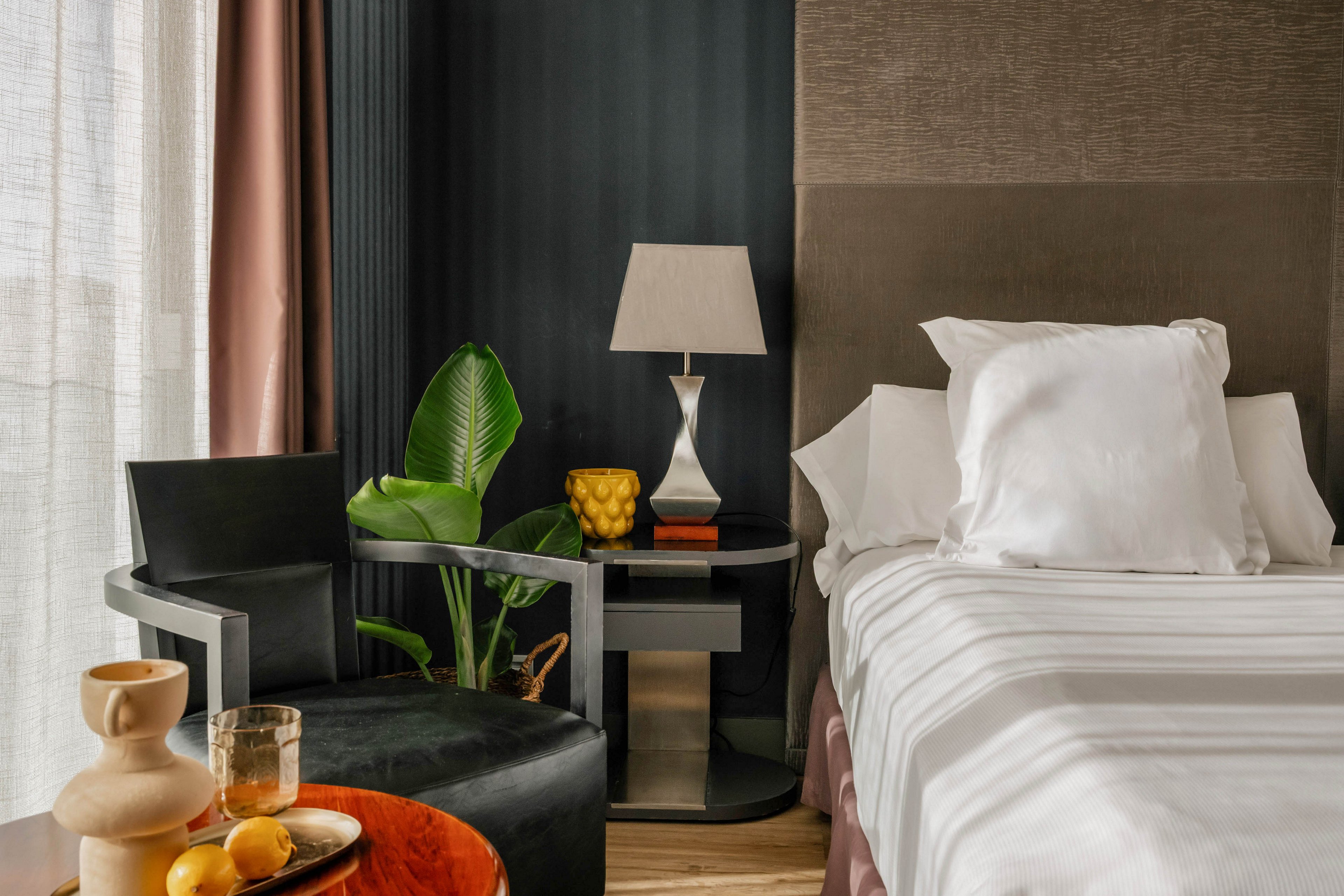 Contemporary bedroom interior featuring a neatly made bed with white linens and pillows. A modern armchair and side table with a sculptural lamp sit beside the bed. A potted green plant, decorative objects, and a bowl of lemons add color and texture to the scene. Soft natural light filters through sheer curtains, creating a warm and inviting atmosphere.