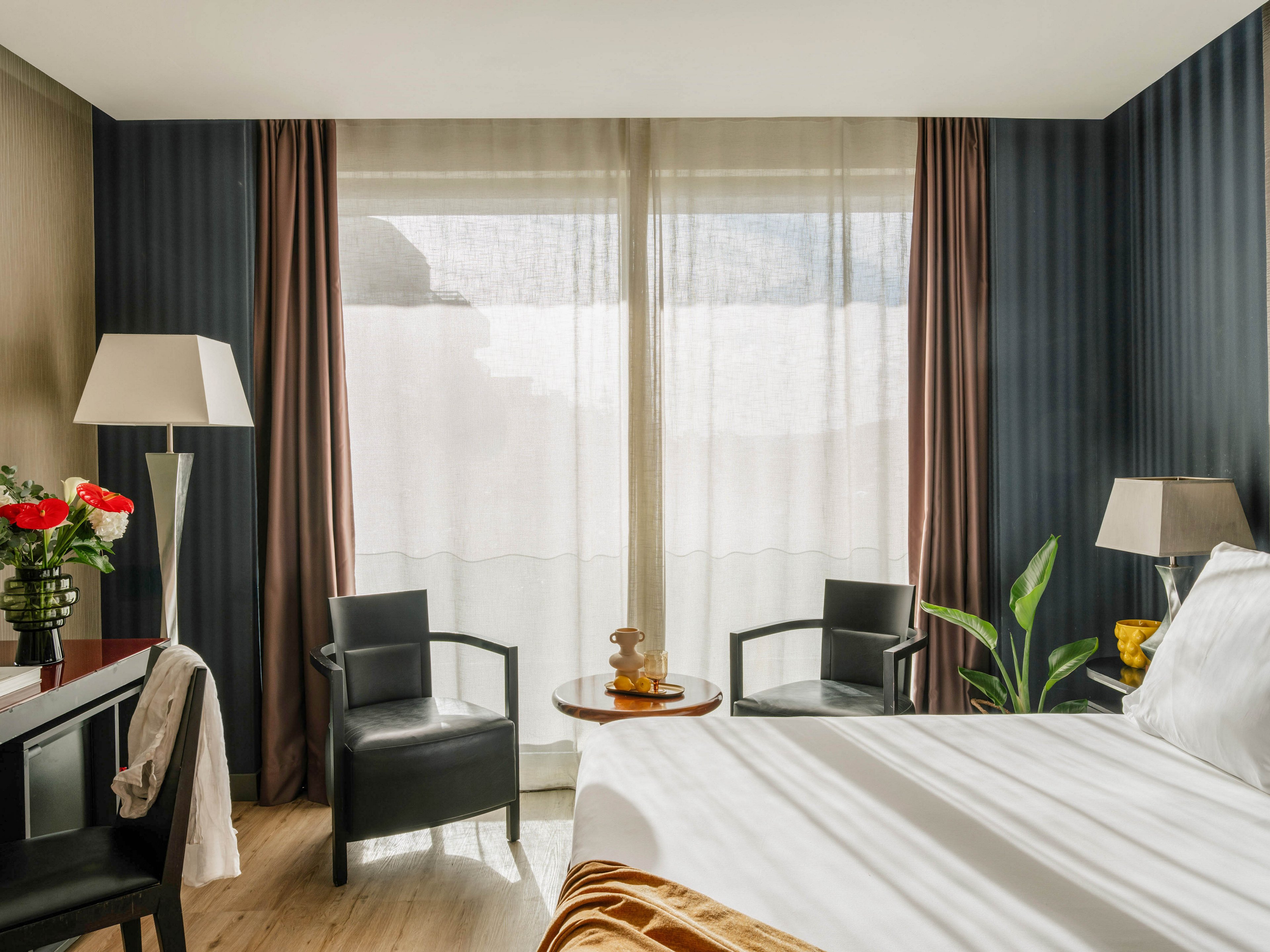 A contemporary hotel bedroom features a neatly made bed with white linens and a golden throw. Two black armchairs and a small round table sit near a large window with sheer curtains and brown drapes, allowing natural light to fill the room. A floor lamp, table lamp, and potted plant add warmth and comfort to the modern decor. Red flowers on a side table and neutral wall tones complete the inviting, stylish atmosphere.