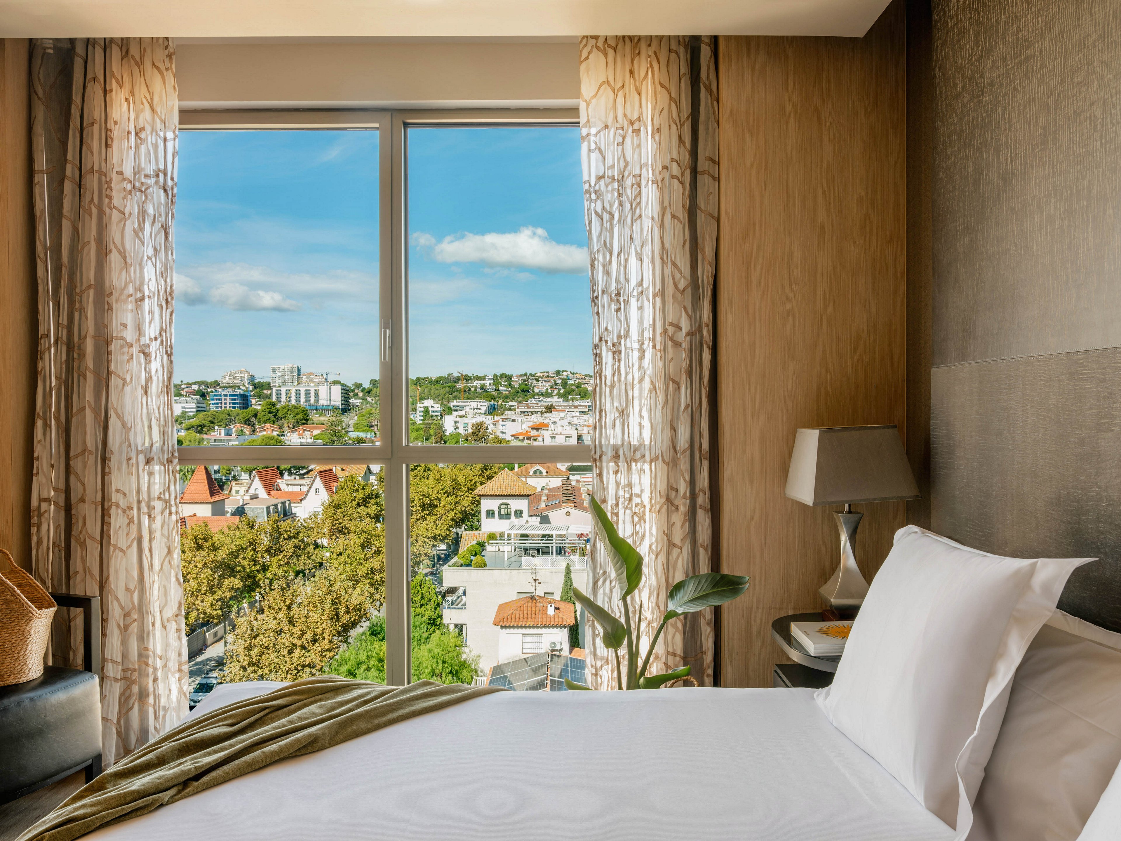A neatly made bed with white linens sits beside a large window in a modern hotel bedroom. Sheer patterned curtains frame the expansive view of a sunny cityscape with trees and low-rise buildings. A bedside table with a lamp and a green plant add warmth and comfort to the interior. The overall scene feels calm, inviting, and filled with natural daylight.