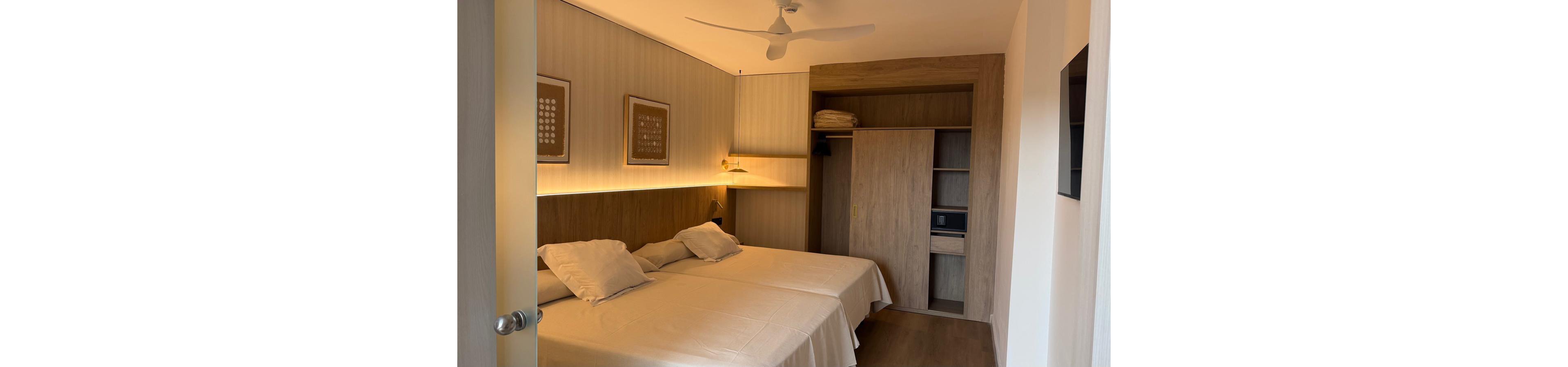 A modern hotel room features two neatly made twin beds with light-colored bedding and pillows. Warm recessed lighting runs along the wooden headboard wall, creating a cozy atmosphere. On the right side, an open wardrobe with shelves, hanging space, and folded towels is visible. Minimalist framed artwork and a ceiling fan complete the clean, contemporary design.