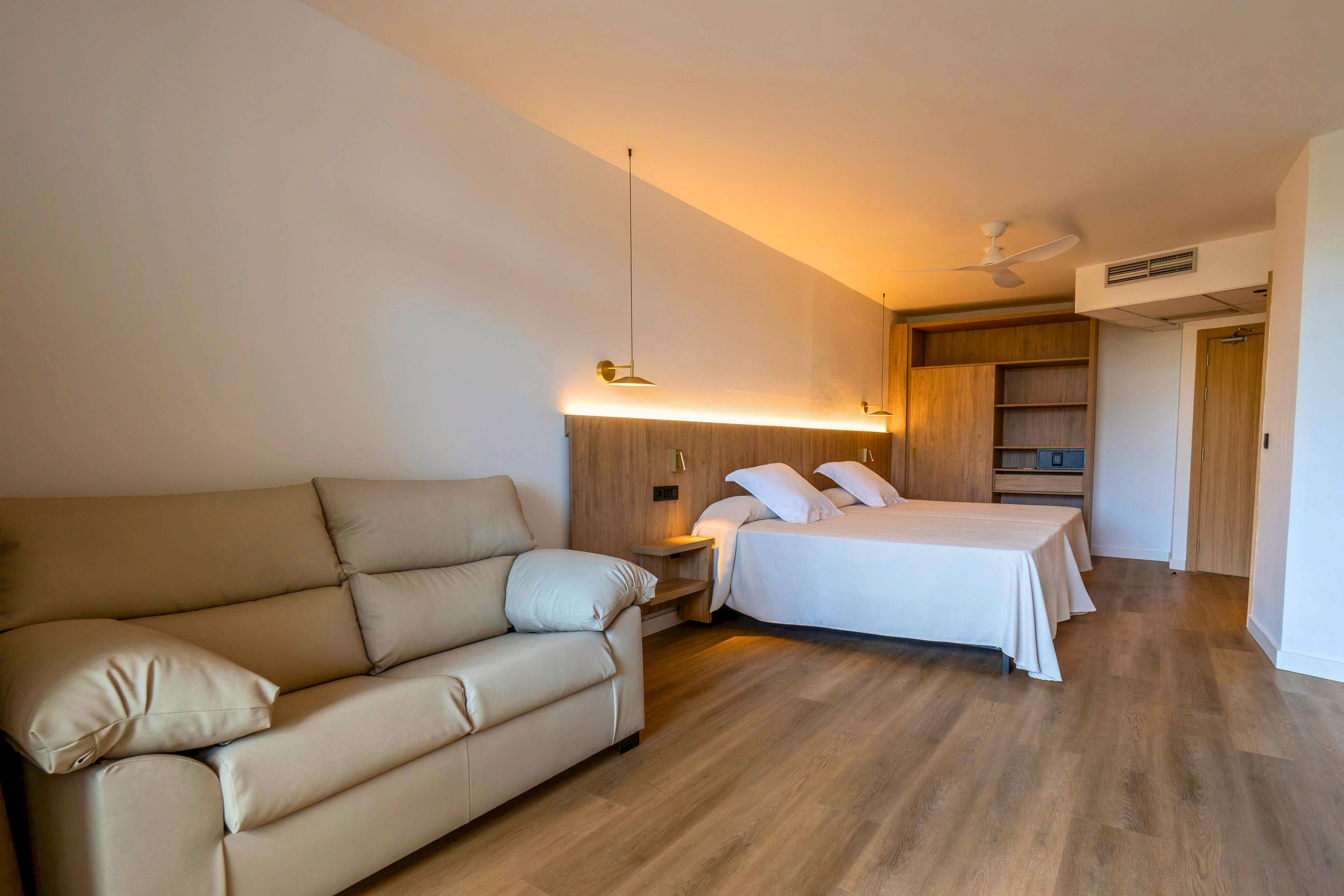 A contemporary hotel room features two neatly made twin beds against a wooden headboard. A beige leather sofa sits along the opposite wall, providing additional seating in the spacious interior. Warm wood flooring and soft ambient lighting create a cozy, minimalist atmosphere. Built-in shelving, a wardrobe, and a ceiling fan complete the clean, functional design.