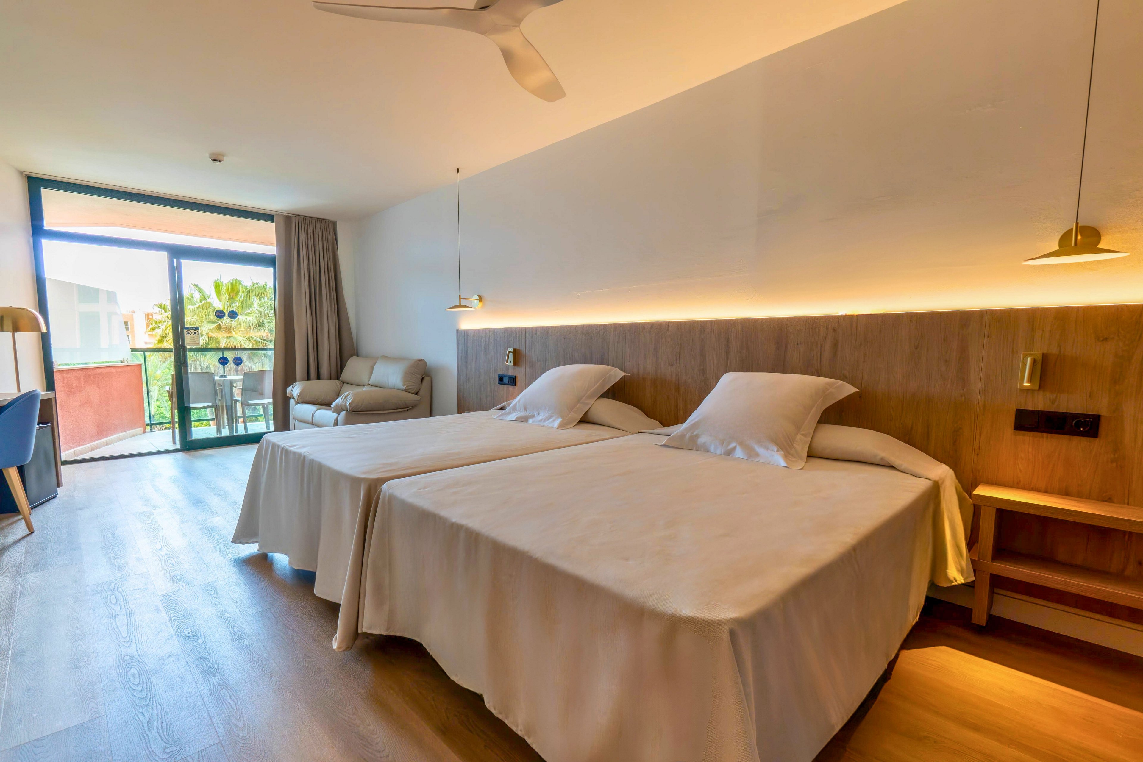 A bright modern hotel room features two neatly made twin beds with light-colored bedspreads. The space includes a sofa near a large glass door that opens to a balcony with outdoor seating. Warm indirect lighting runs along the wooden headboard, complemented by minimalist pendant lamps and a ceiling fan. Natural light fills the room, highlighting the clean lines and contemporary decor.