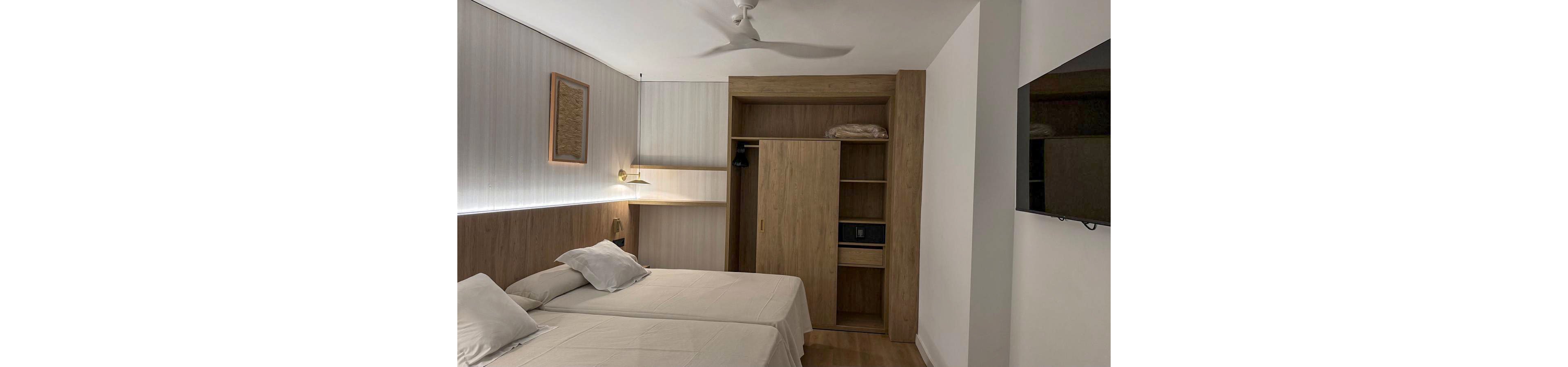 A contemporary hotel room features two neatly made twin beds with white linens and pillows. The space includes a built-in wooden wardrobe with open shelves, a safe, and folded bedding. Soft wall lighting and a ceiling fan create a calm, comfortable atmosphere. A flat-screen television is mounted on the opposite wall, completing the minimalist interior design.