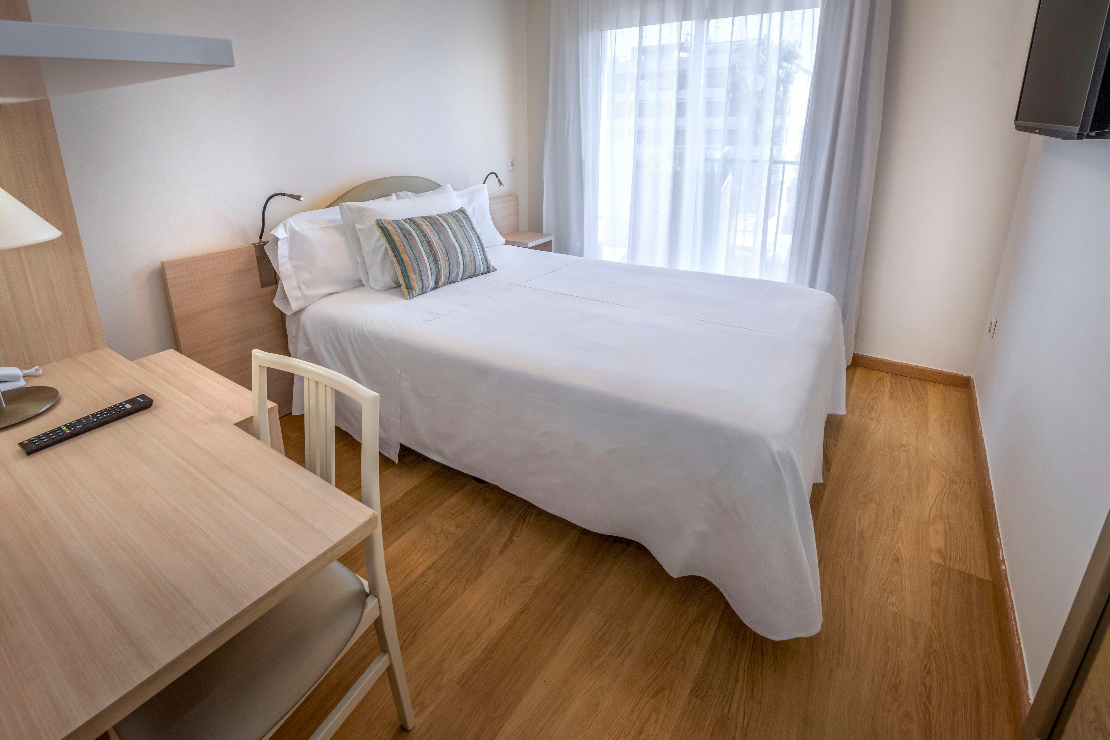 A neatly made double bed with white linens is the focal point of this bright bedroom. The room features light wood furniture, including a small desk with a chair and a television remote placed on top. Sheer curtains cover a large window or balcony door, allowing natural light to softly illuminate the space. The overall design is simple and modern, creating a clean and inviting atmosphere.